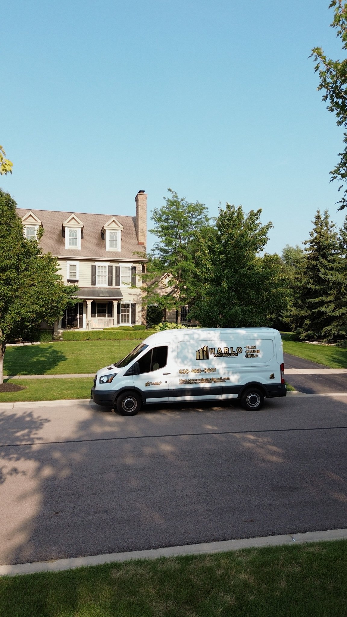 A white construction company van parked on the side of a suburban street in front of a large house with green lawns and trees.