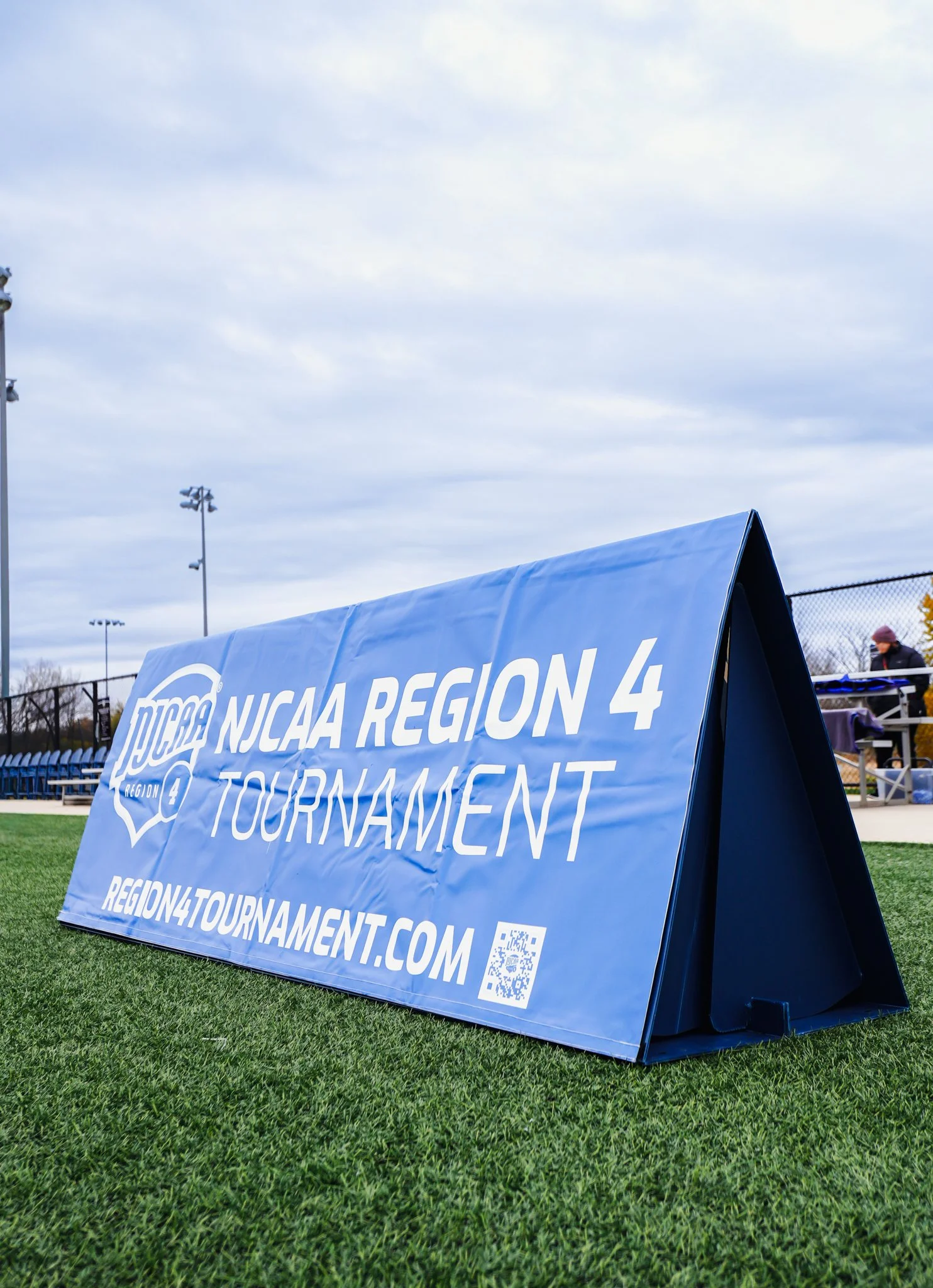Blue sign on grass reads 'NJCAA Region 4 Tournament' with website 'REGION4TOURNAMENT.COM' and a QR code. In the background, there are stadium lights, a chain-link fence, and some people sitting on bleachers under a cloudy sky.