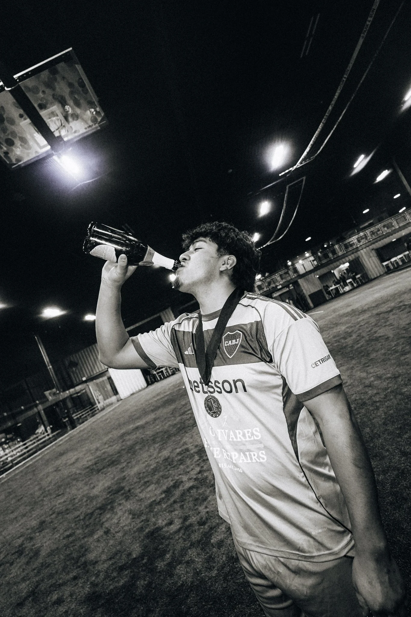A young man with curly hair wearing a sports jersey drinks from a bottle while standing on a soccer field, wearing a medal around his neck.