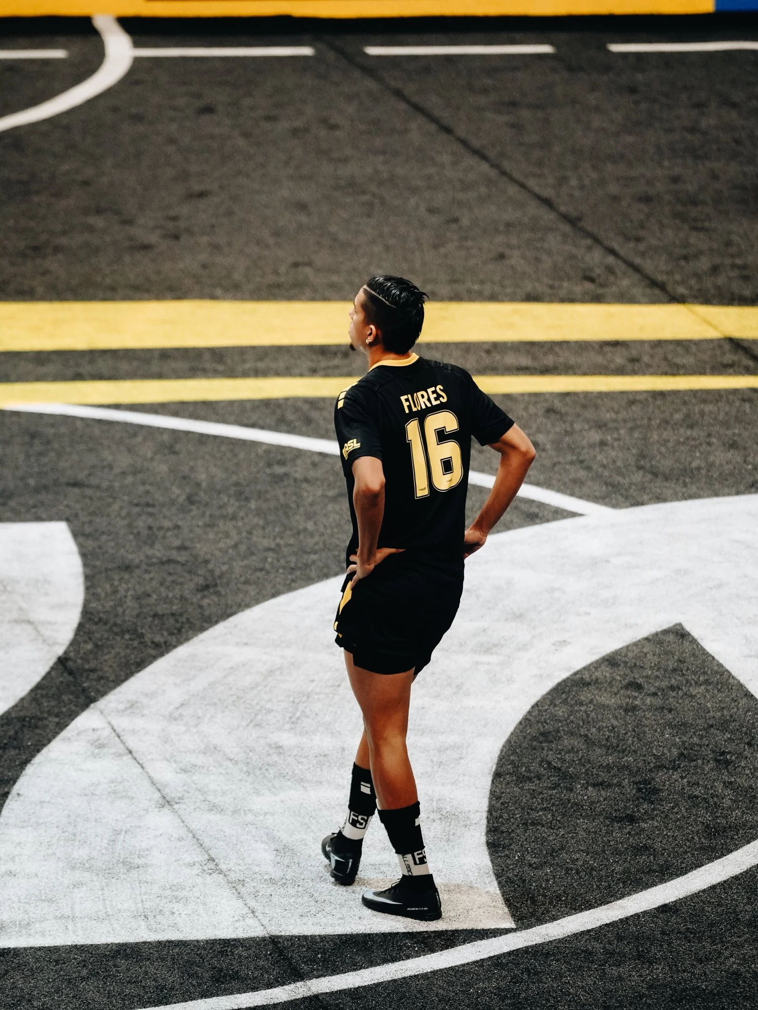 A soccer player wearing a black jersey with the number 16 and the name 'Flores' stands on a track at a sports stadium.