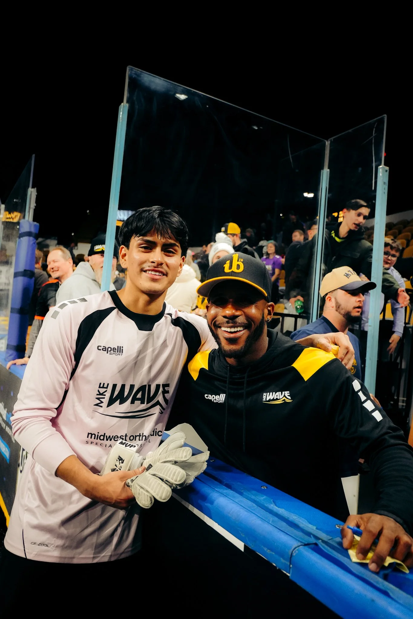 Two men smiling, one in a white goalie jersey and gloves, and the other in a black jacket with yellow accents, standing together at a sports event, with a crowd in the background.