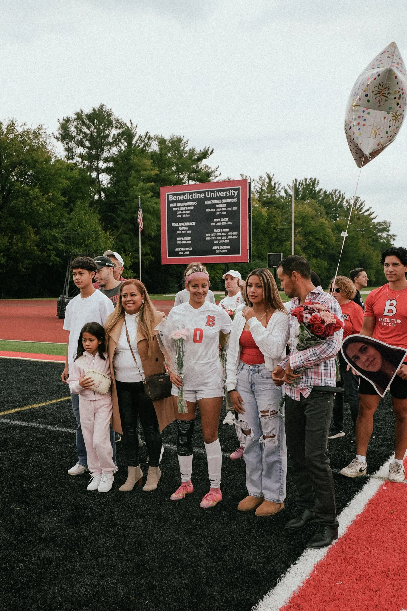 Group of people on a sports field celebrating, with a girl in soccer uniform holding pink flowers, surrounded by family and friends, in front of a Benedictine University sign and a balloon.