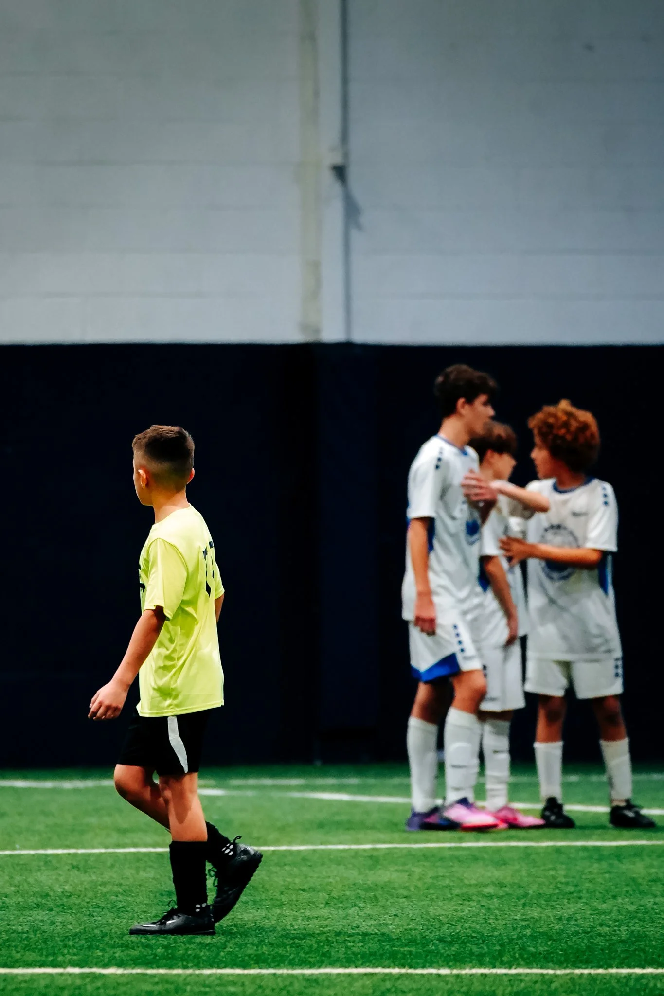 Young soccer players in uniform on an indoor field, with one boy standing alone in the foreground and a group of three boys conversing in the background.
