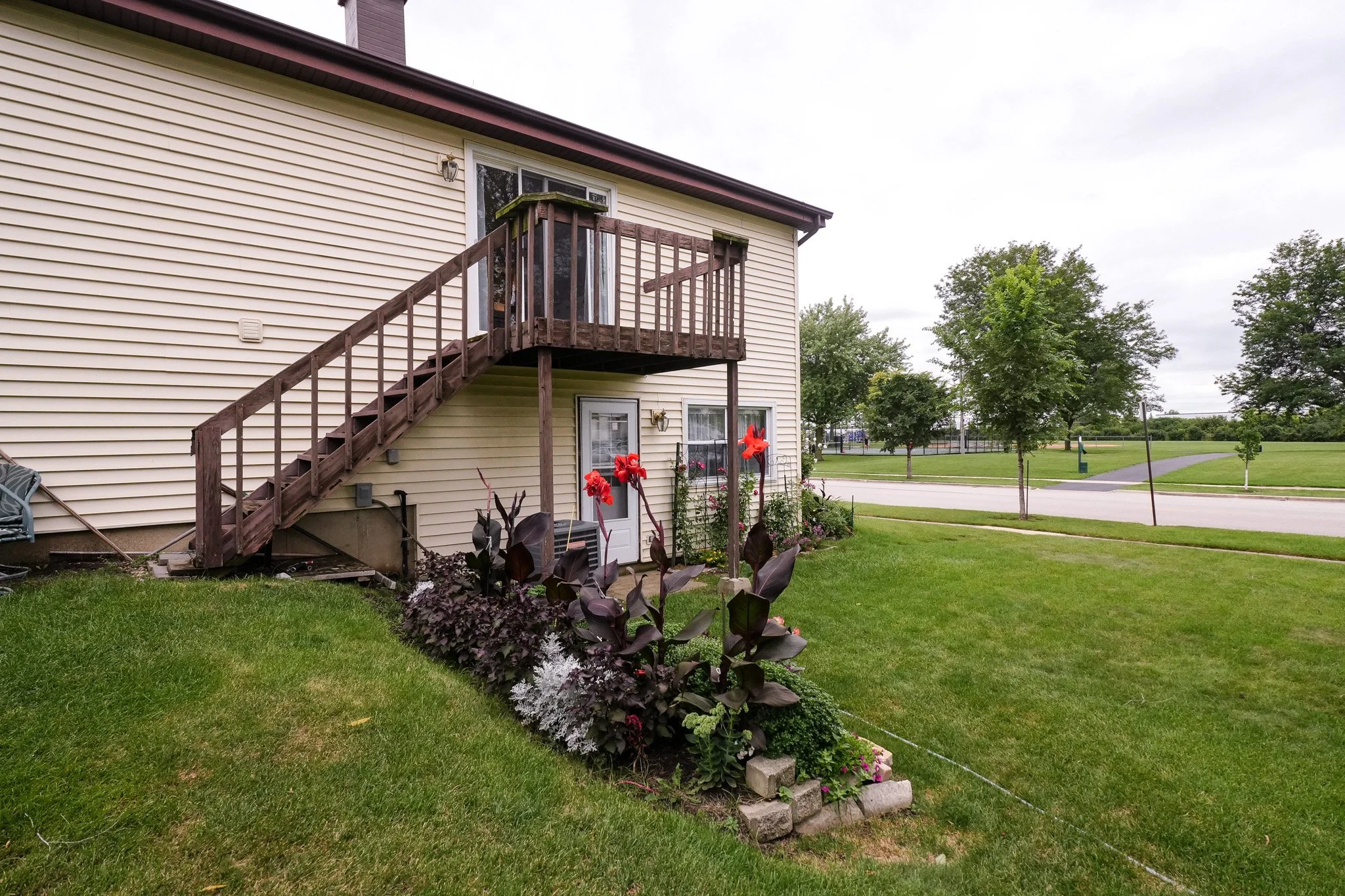 Backyard view of a two-story house with a wooden staircase leading to a second-floor deck. There are flower beds with red lilies and dark purple foliage plants in the yard, alongside a well-maintained lawn and a park with trees and a walking path in the background.