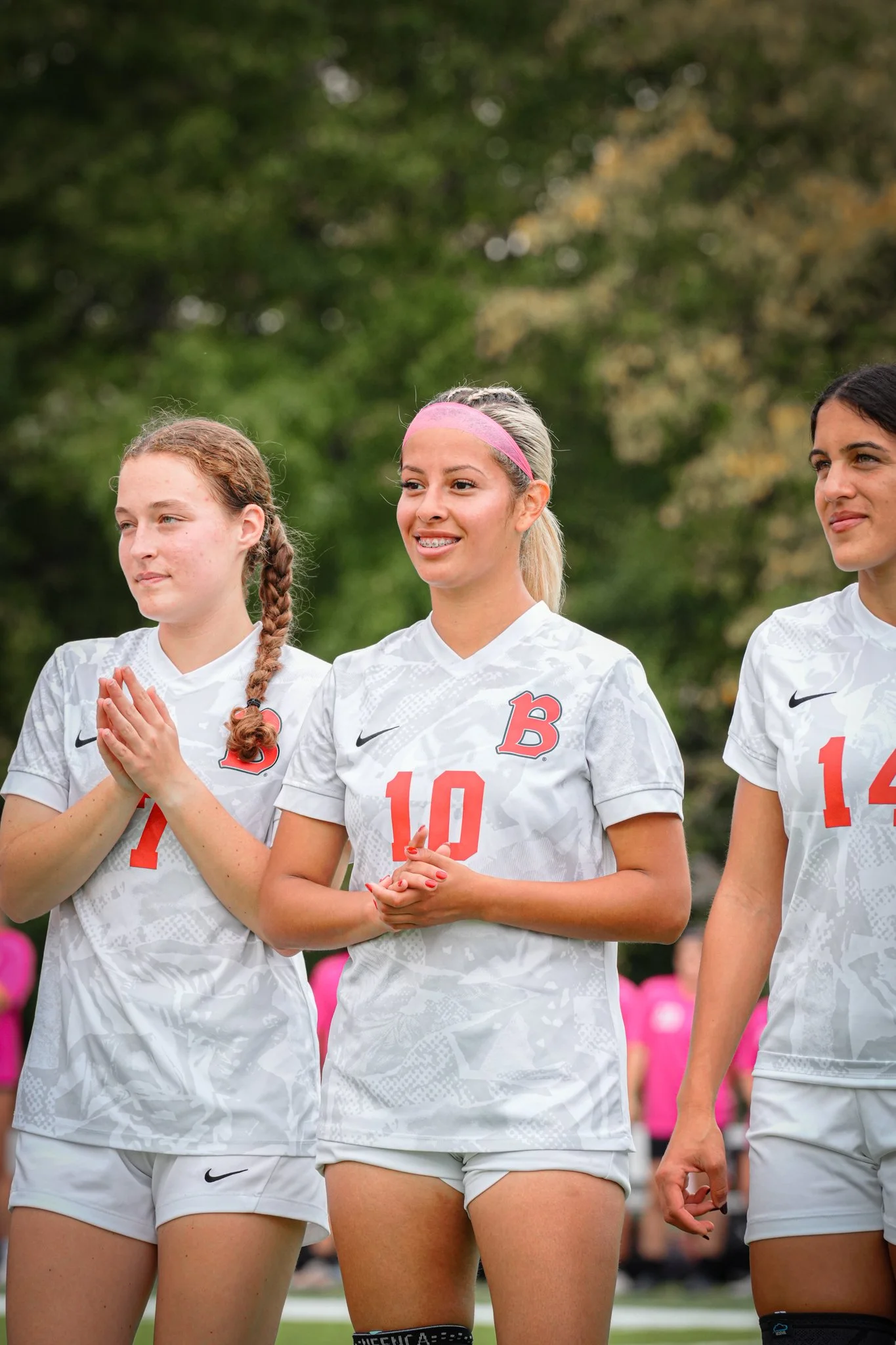 Three female soccer players standing together outdoors, wearing white jerseys with red numbers and logos, during a game or event.