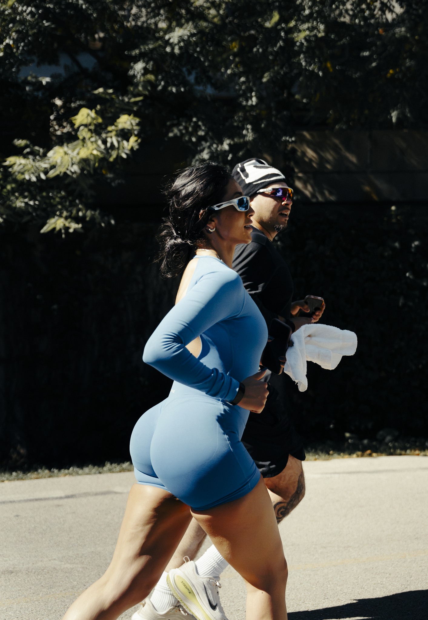 A woman in a blue athletic outfit running outdoors with a man in black sportwear behind her, both wearing sunglasses and headphones.