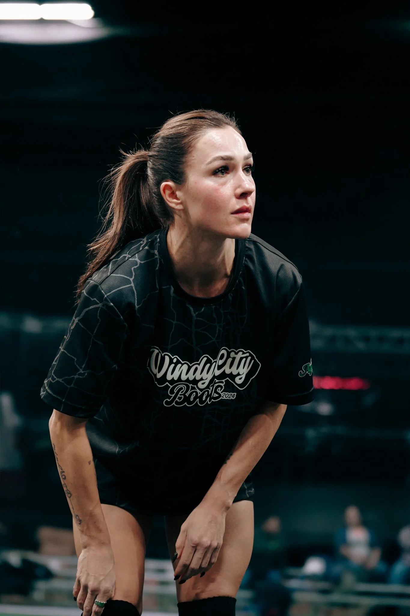 Female athlete in black 'Windy City Bods' shirt, leaning forward, in an indoor sports facility, with people and vehicles blurred in the background.