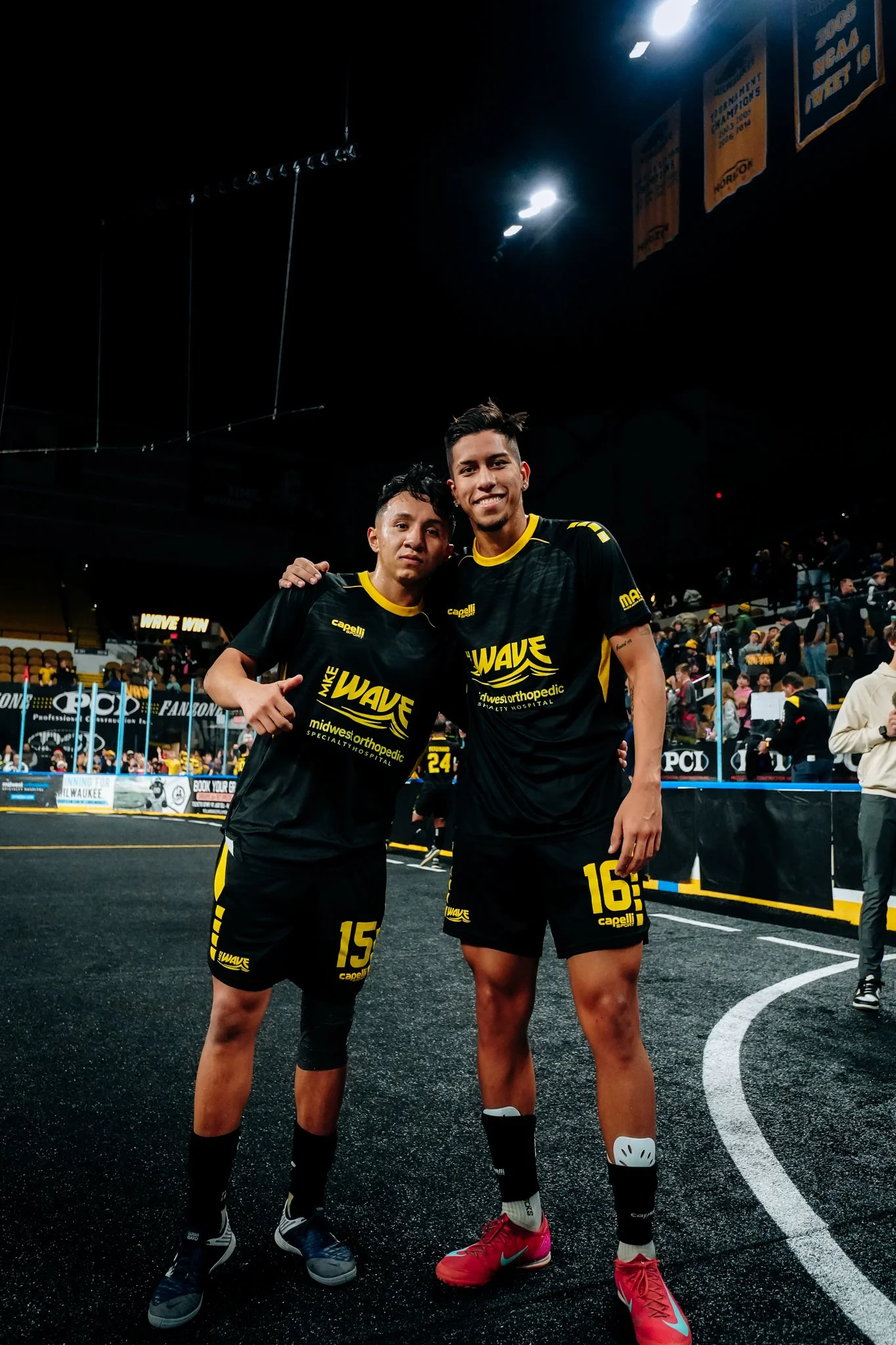 Two male athletes in black and yellow sports uniforms posing together after a game, on an indoor field with a crowd in the background.
