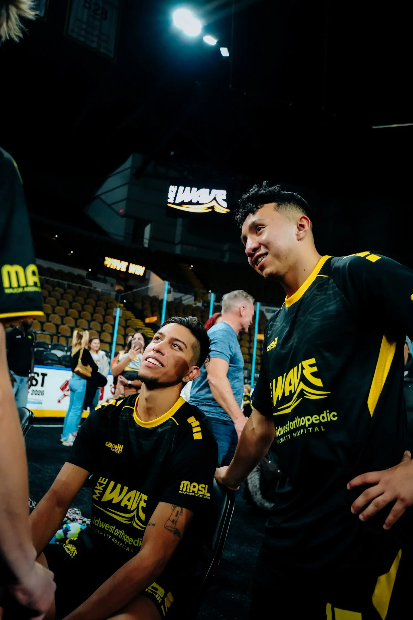 Two basketball players in black and yellow uniforms sitting and standing in an arena, smiling and talking, with a few spectators and a scoreboard in the background.