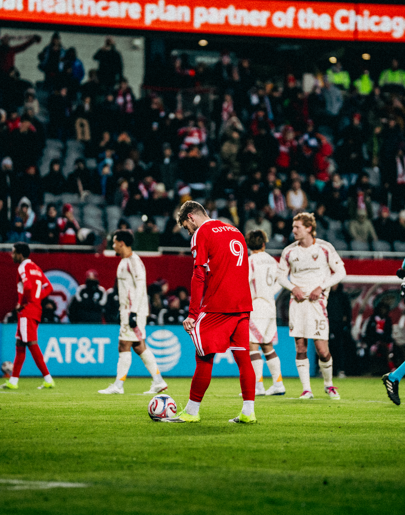 A soccer player wearing a red jersey with the number 9 stands on the field with his head down, next to a ball. Other players in white and red jerseys are visible in the background, along with spectators in the stands.