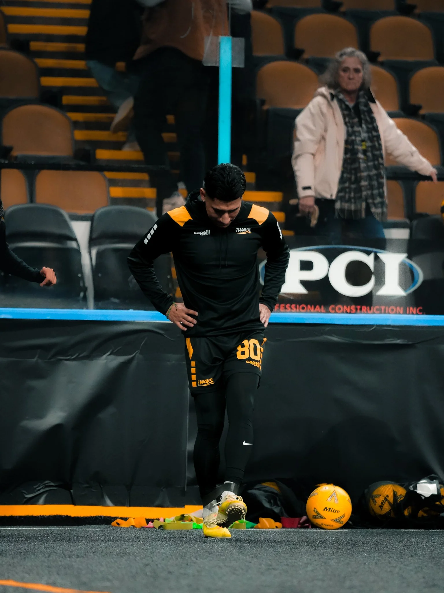 A male athlete in black and yellow sportswear standing on the sidelines of a sports event surrounded by sports equipment, including balls and cones, with spectators in the background.