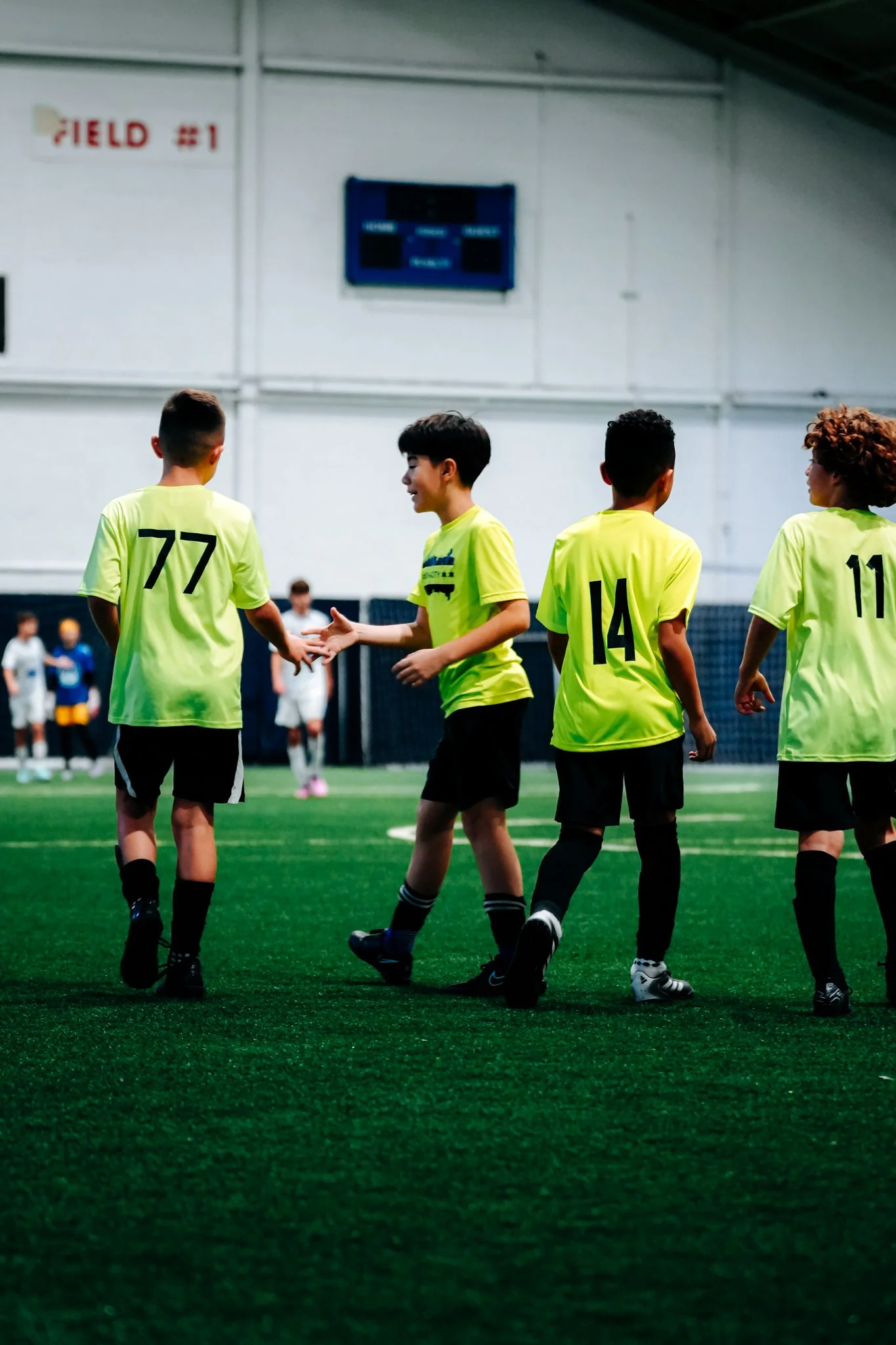 Young boys playing soccer indoor, wearing yellow jerseys numbered 77, 14, and 11, shaking hands after a game.