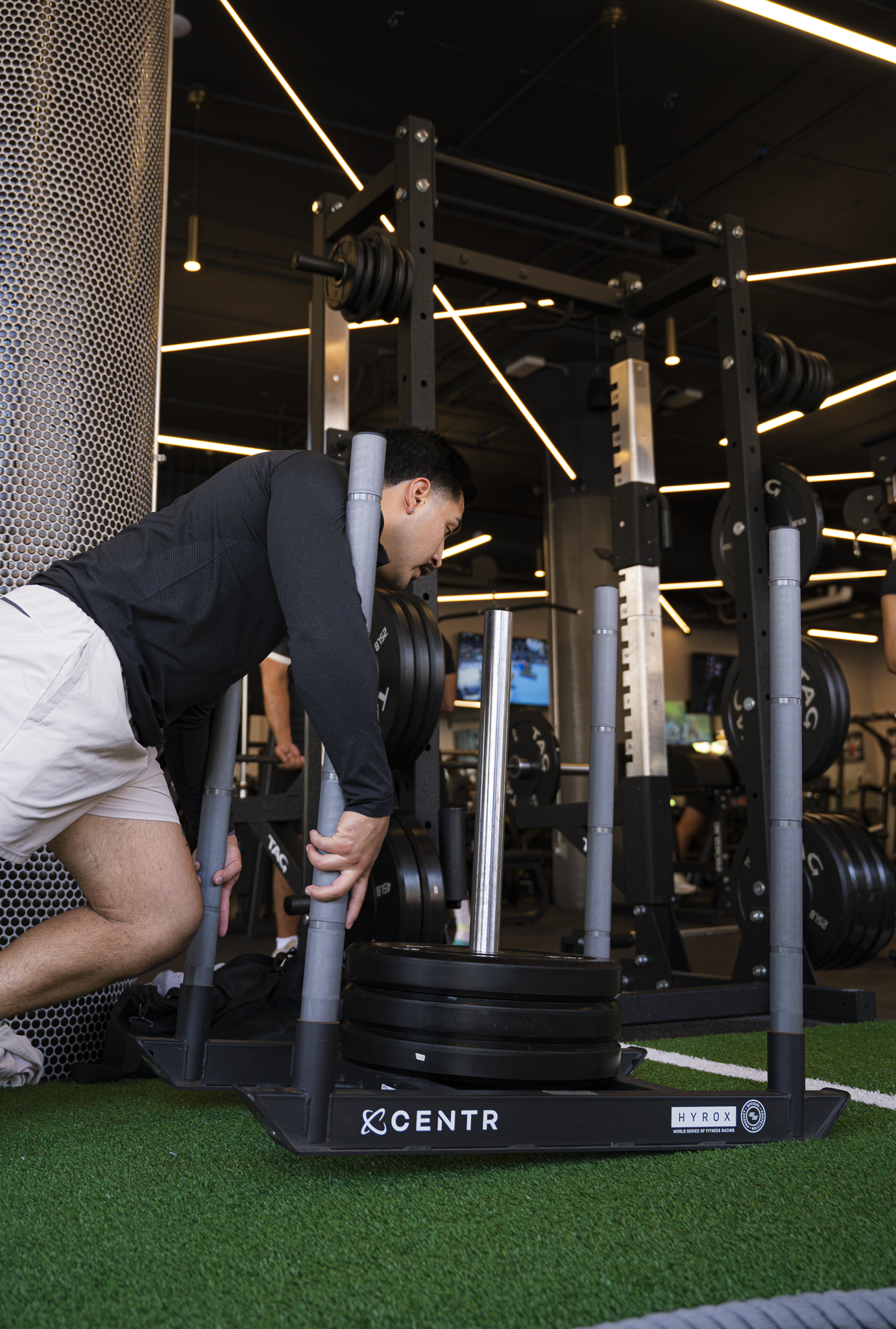 A man wearing a black long-sleeve shirt and white shorts is doing a sled push exercise in a gym. He is pushing a weighted sled on green turf with a modern fitness facility in the background, featuring black ceilings, linear LED lighting, and weight racks.