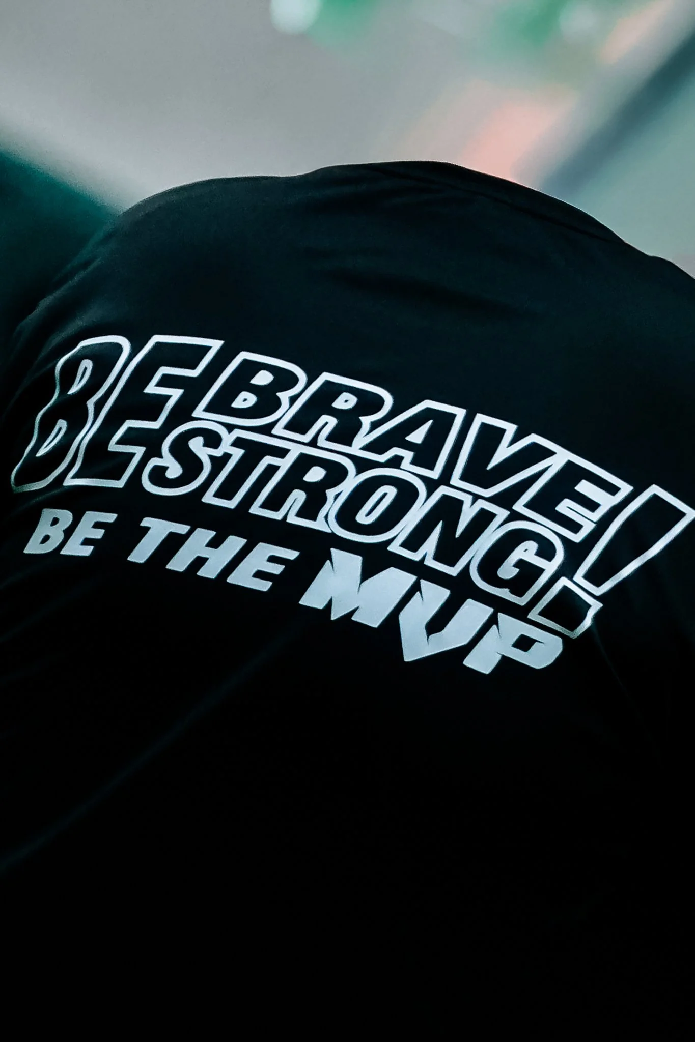 A close-up of a black shirt with white text that reads "BRAVE! BE STRONG! BE THE MVP".