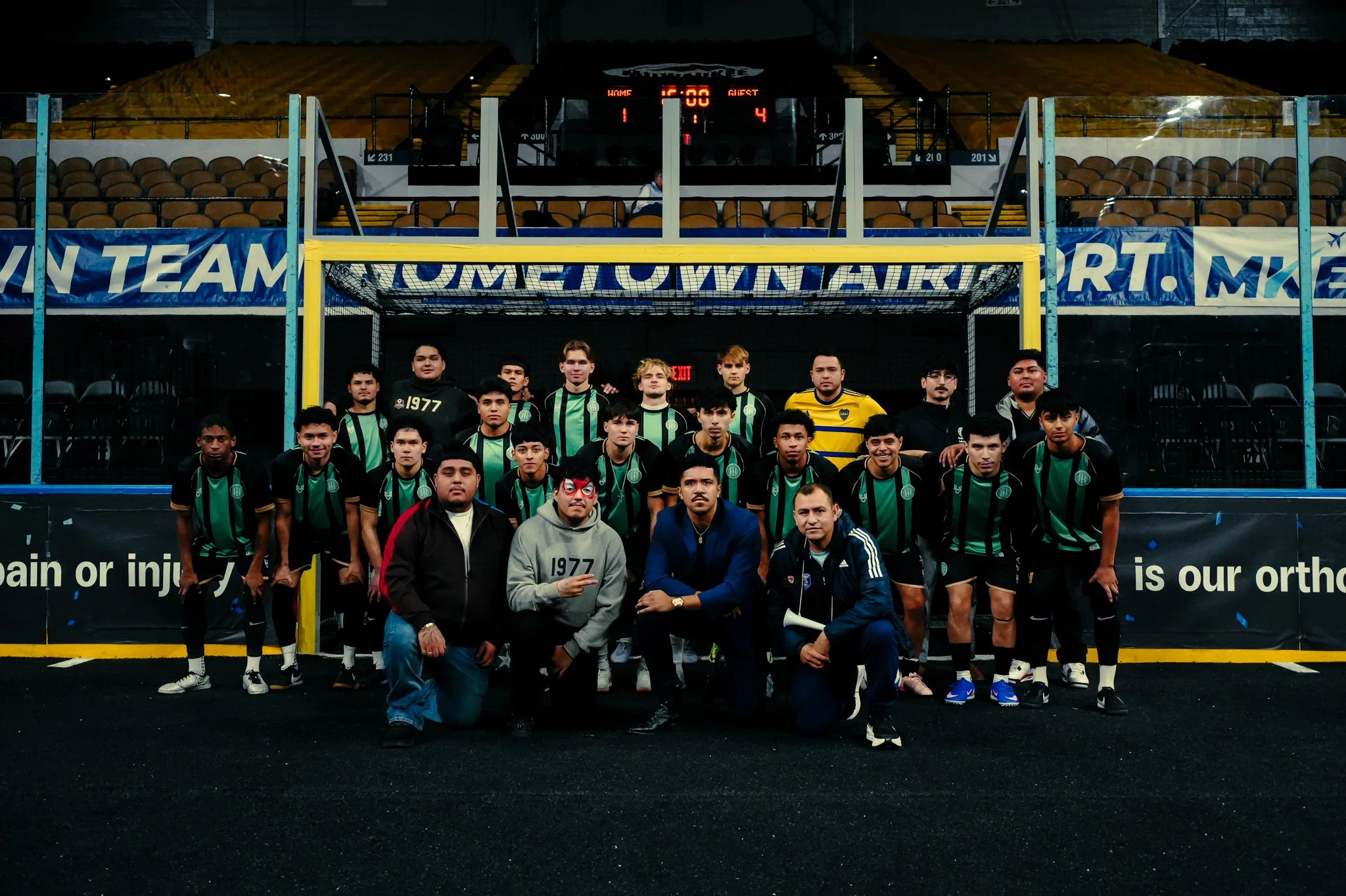 A group of young soccer players and their coaches posing for a team photo on a soccer field at night, with an empty stadium and a scoreboard in the background.