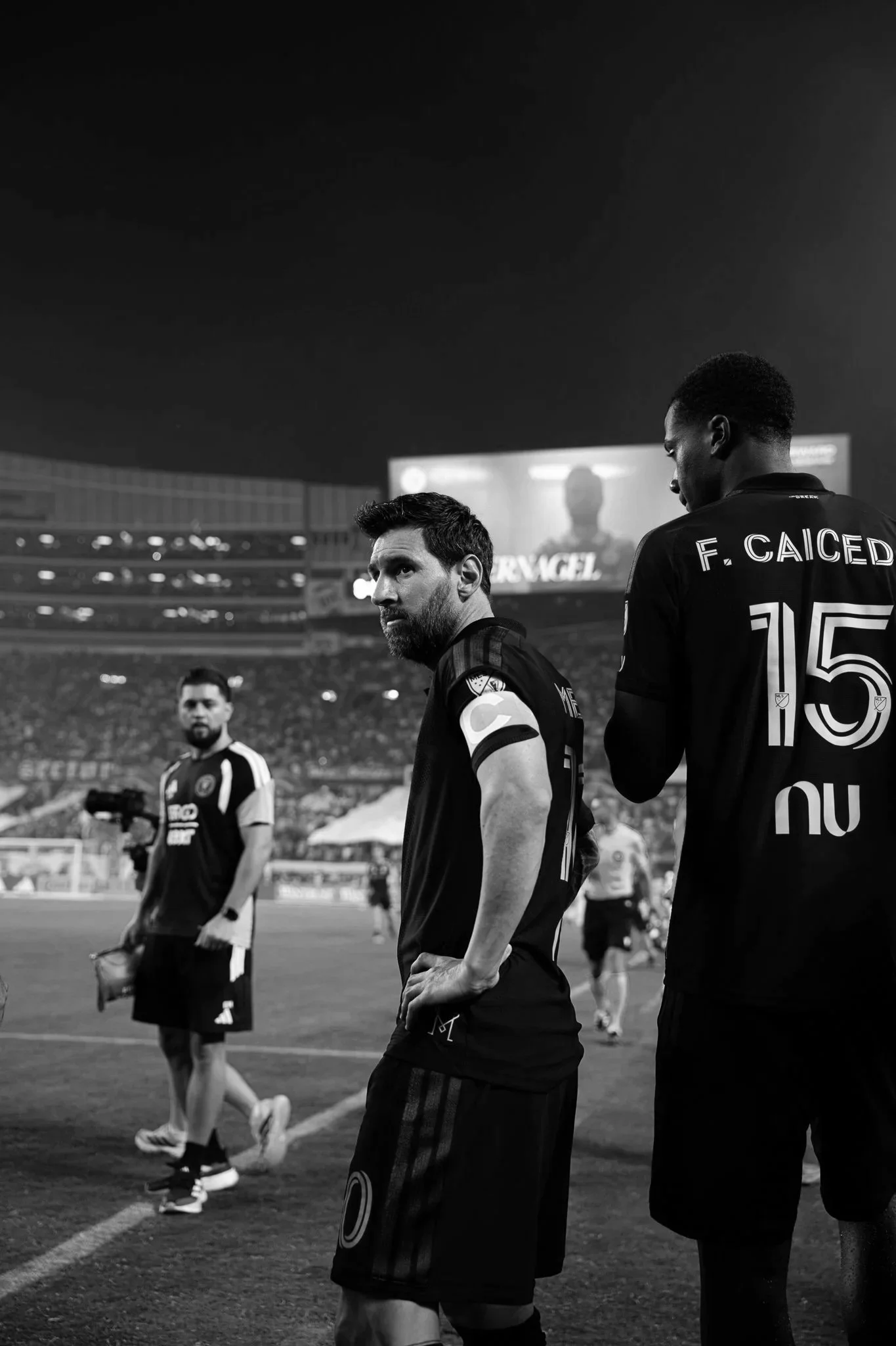Two soccer players standing on the field during a match, with a large screen displaying a person in the background, black and white photo.