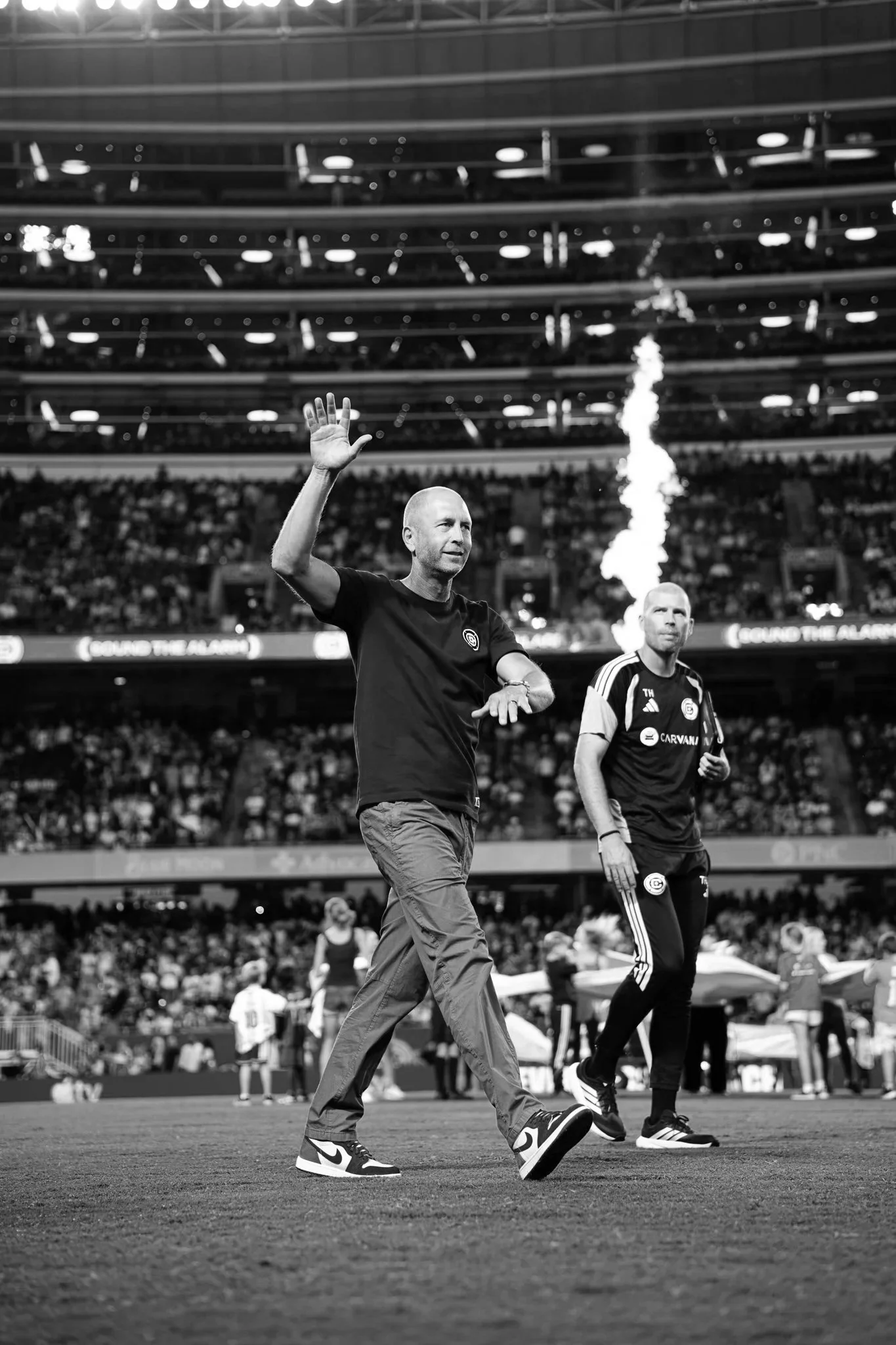 Black and white photo of two men on a soccer field; one man in a black shirt is waving, the other man in a black and white uniform is standing nearby; stadium with multiple levels of seating and spectators in the background.