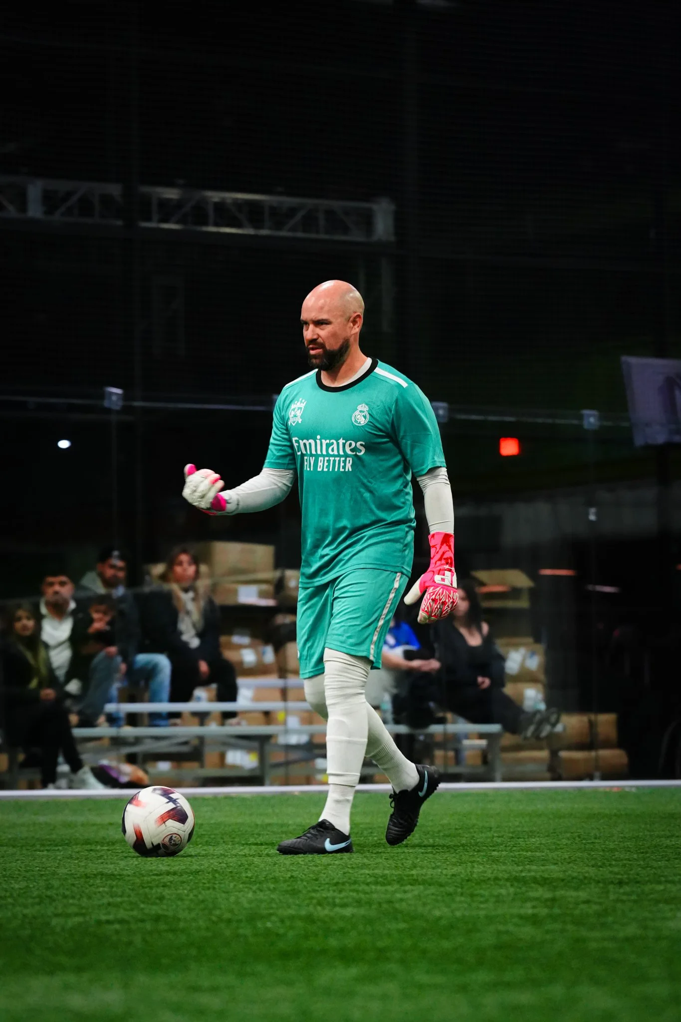 A male soccer goalkeeper in a teal jersey and white goalie gear on a soccer field, with spectators in the background.
