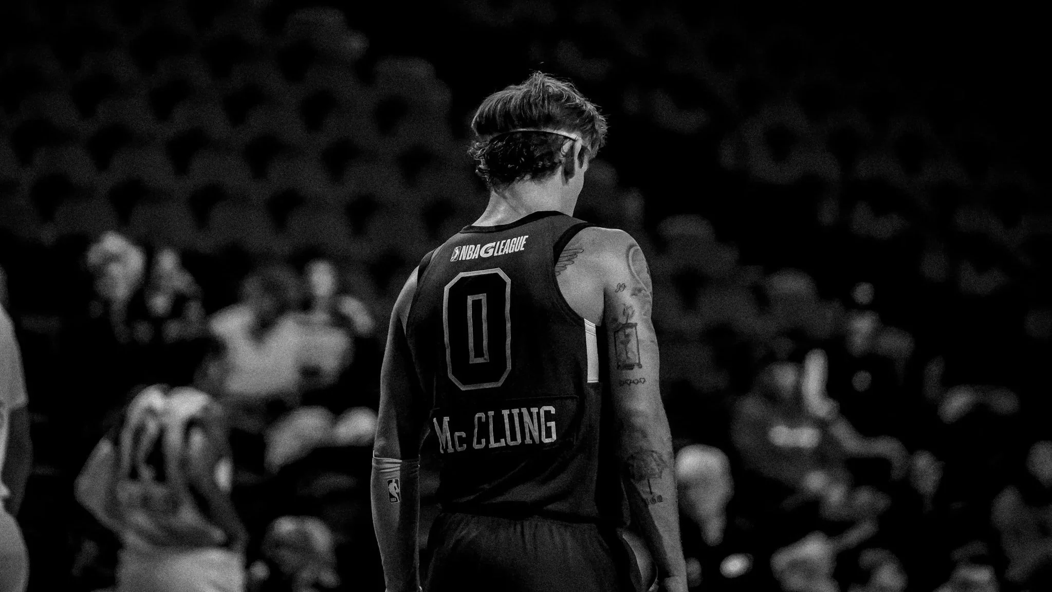 A basketball player with tattoos, wearing a jersey with the name 'Mc CLUNG' and the number 0, stands on the court with his back facing the camera, in a dimly lit environment with spectators in the background.