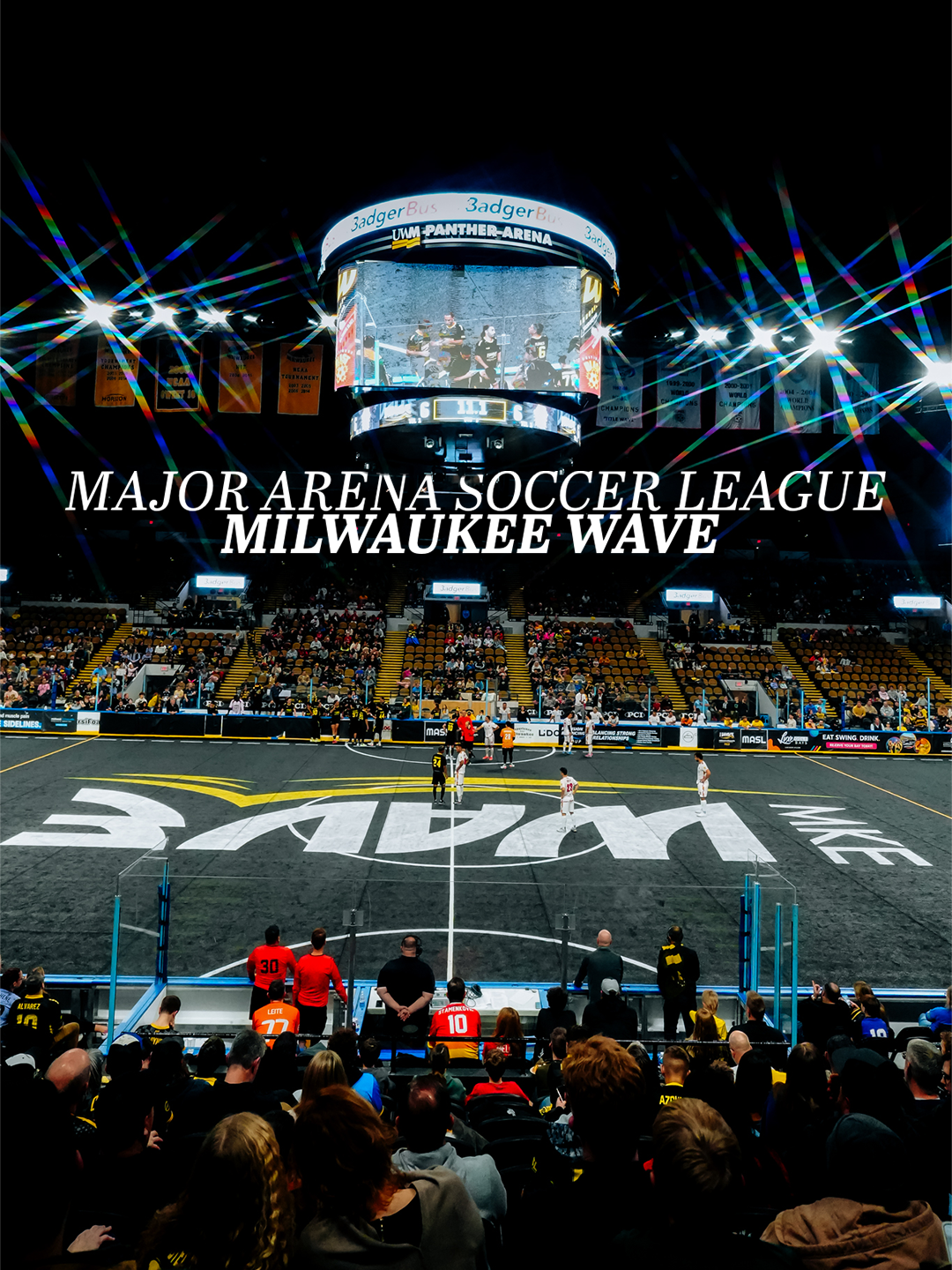 Indoor arena with a black floor and soccer markings, spectators seated in front, a scoreboard displaying players, and banners hanging in the ceiling. Text overlays indicate "Major Arena Soccer League Milwaukee Wave."