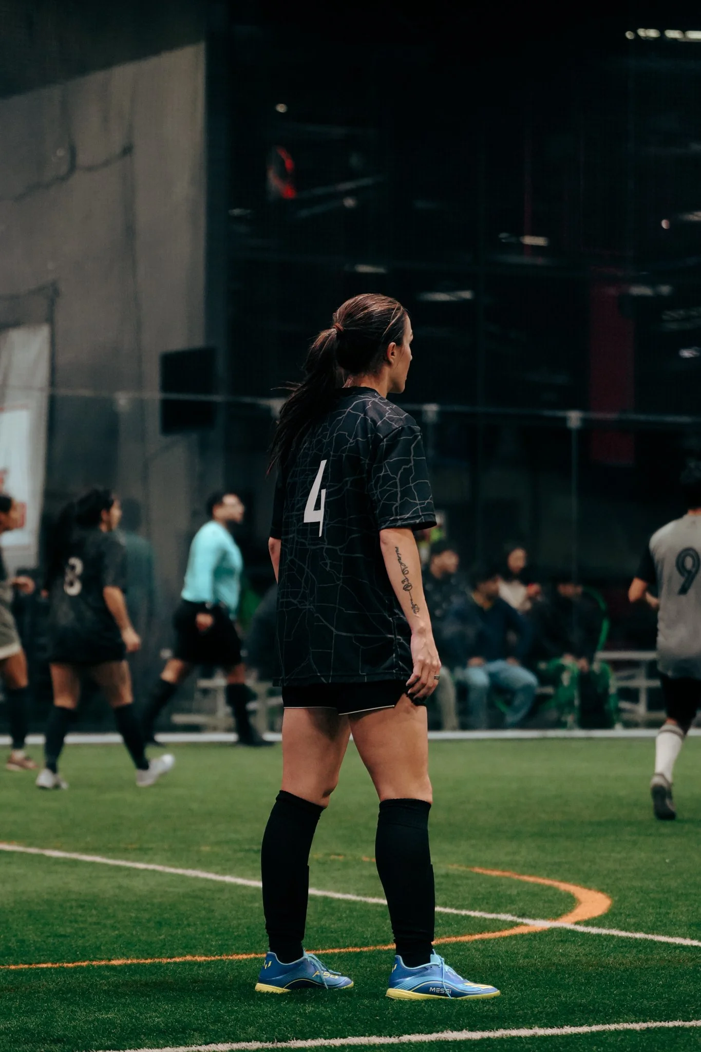 A female soccer player in a black jersey with the number 4 stands on the field, watching the game as other players and spectators are visible in the background.