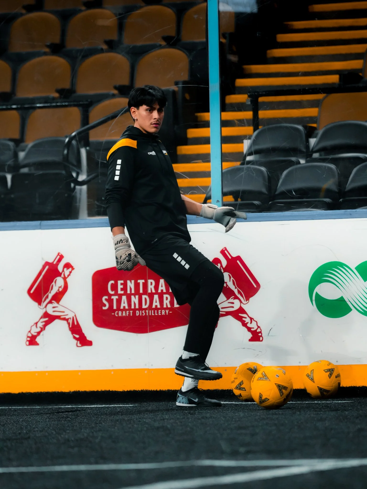 Hockey player in black uniform standing on ice rink, holding hockey stick, with three yellow hockey balls on the rink floor, in front of advertising boards with logos and empty seating area in the background.
