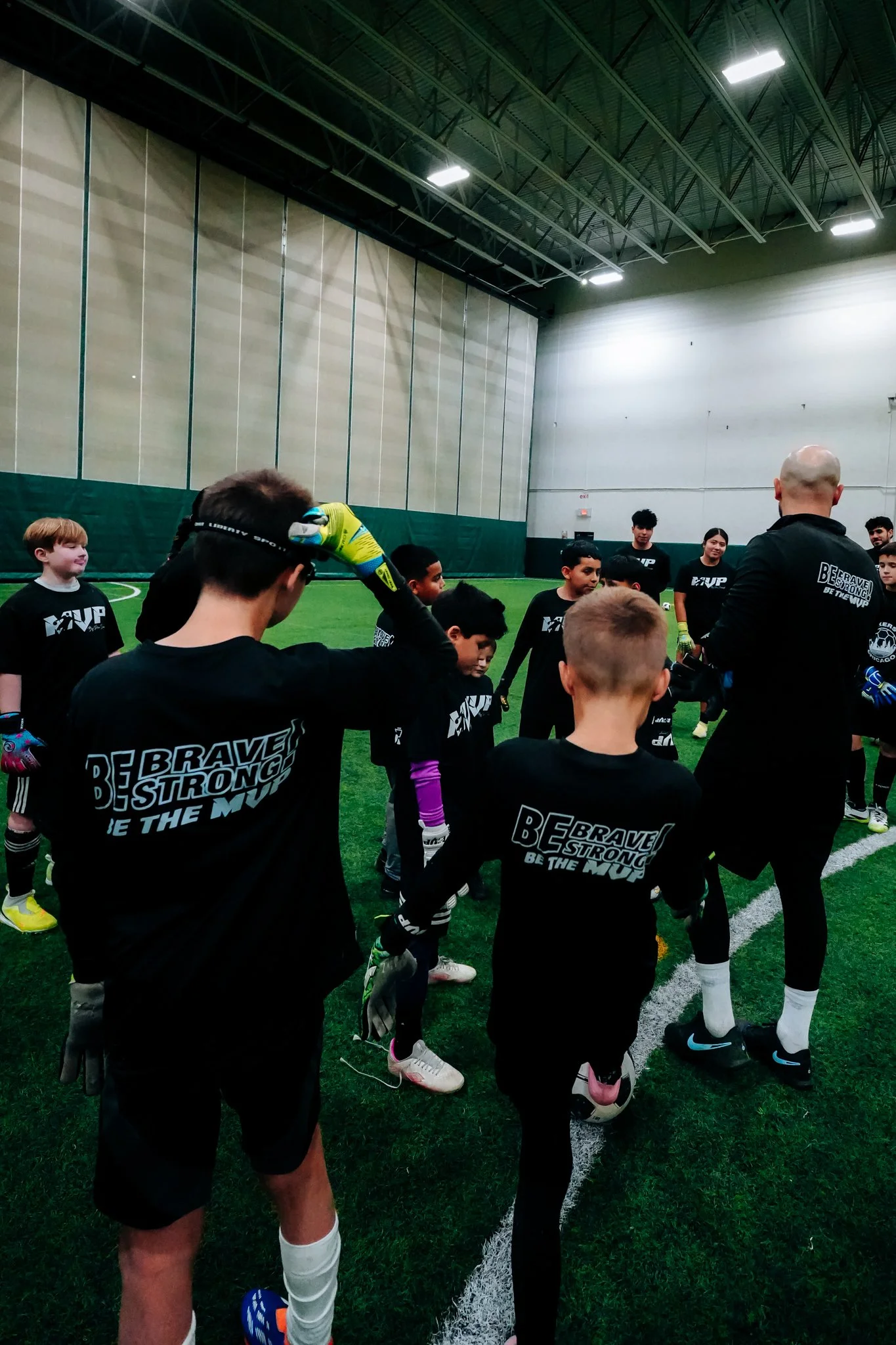 A group of young soccer players and their coach inside an indoor soccer facility, standing on the field in a semi-circle around the coach.