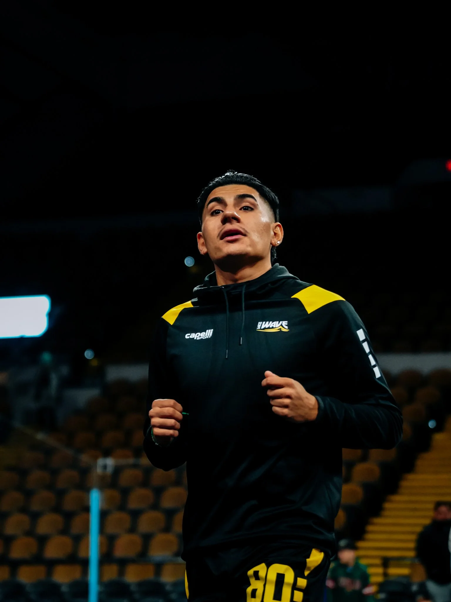 A man jogging indoors, wearing a black athletic jacket with yellow accents, black pants with yellow pattern, and a focused expression.