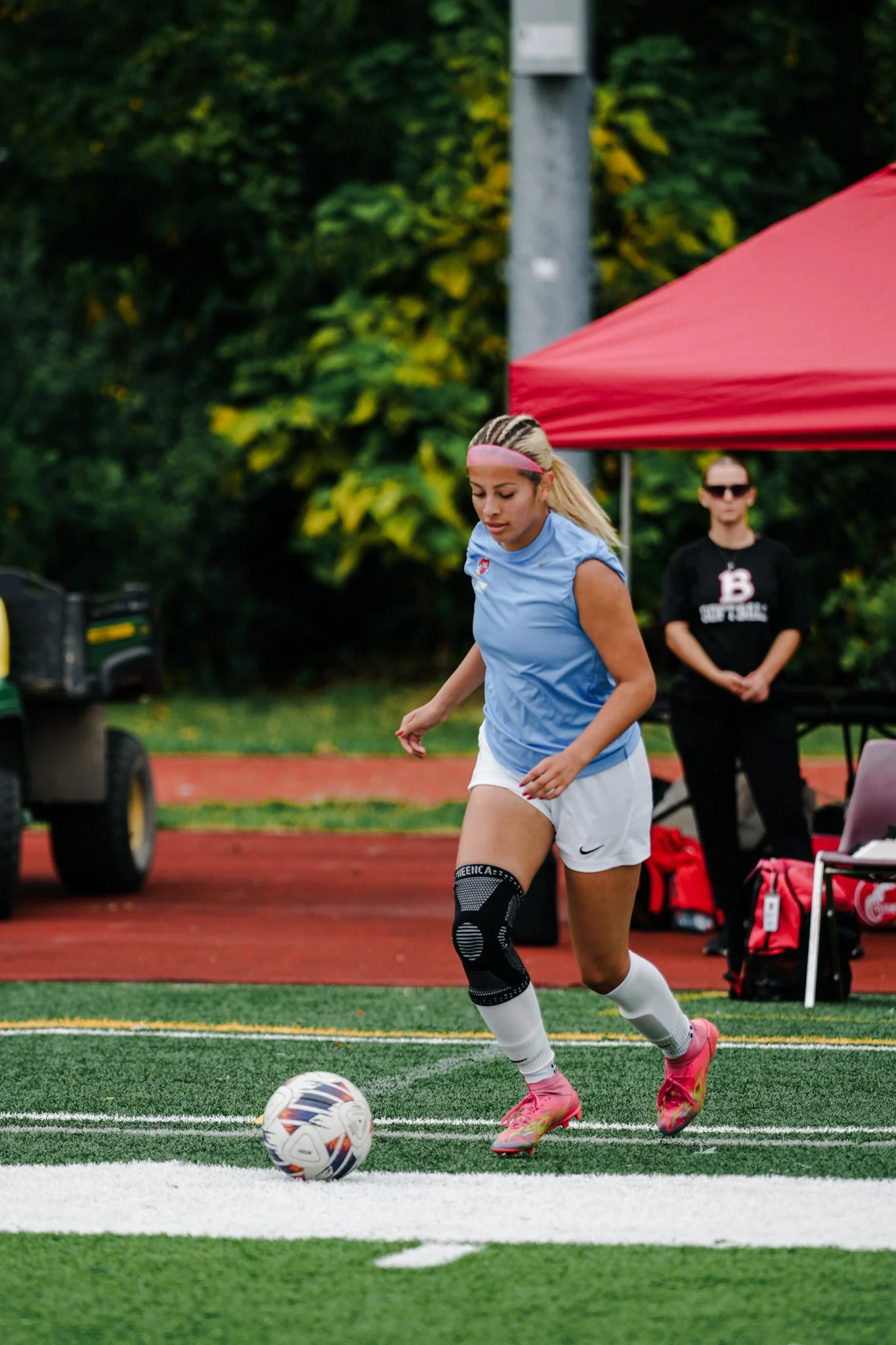 A female soccer player in a blue jersey and white shorts is about to kick a soccer ball on a turf field. She is wearing pink cleats and a black knee brace on her right leg. A red canopy and a woman in black clothing and sunglasses are in the background.