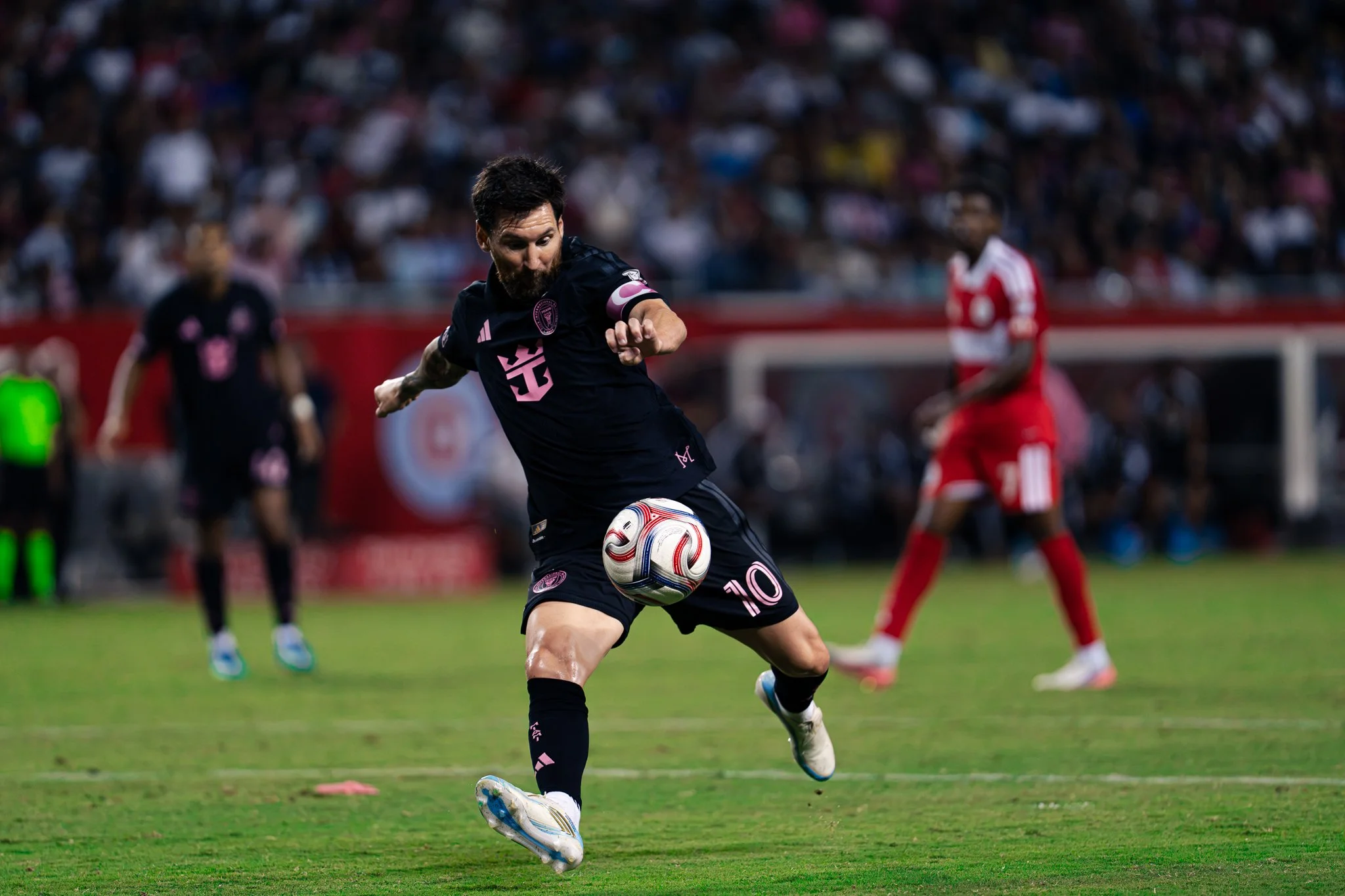 A male soccer player with a beard and dark hair, wearing a black jersey with pink accents and the number 10, is kicking a white soccer ball on a green field during a match. There are other players and fans in the background.