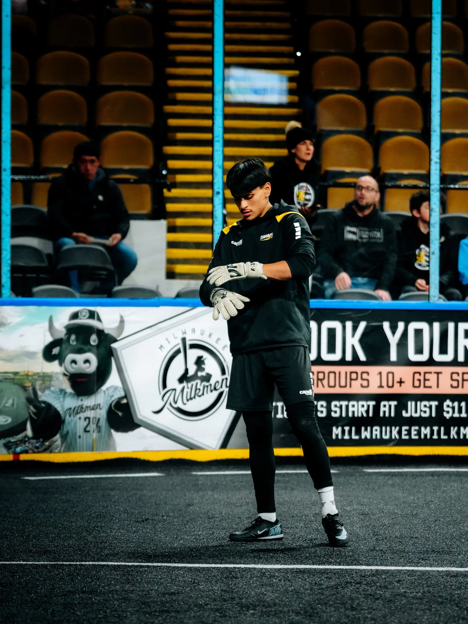 A young soccer player wearing black athletic gear, including gloves, stands on a turf field. He appears to be focused, with arms crossed, in front of a stadium barrier with advertisements and spectators sitting in the background.