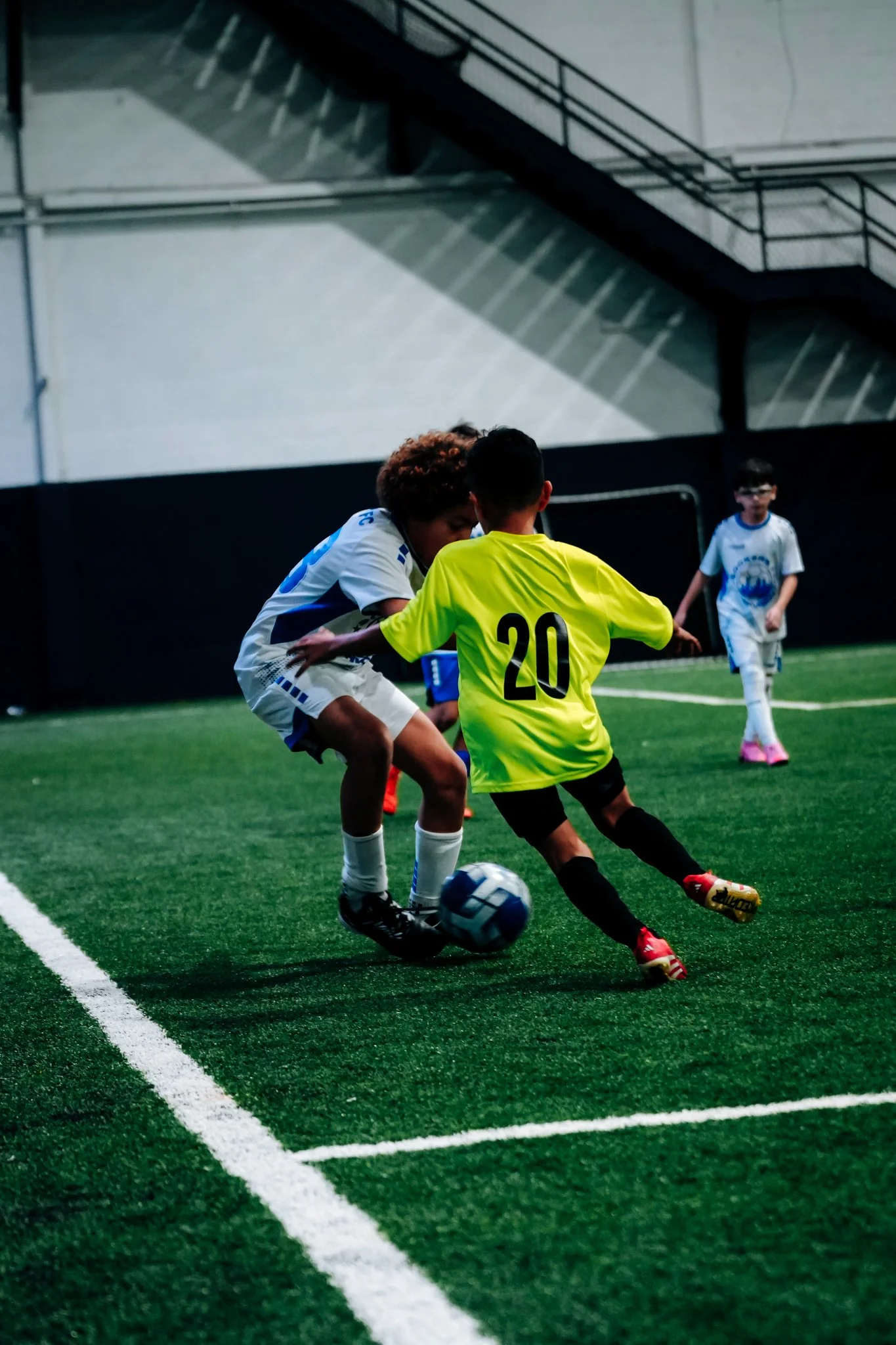Children playing soccer indoors, two kids in the foreground battling for the ball, one in a white uniform and another in a yellow jersey with the number 20, with additional kids in the background.