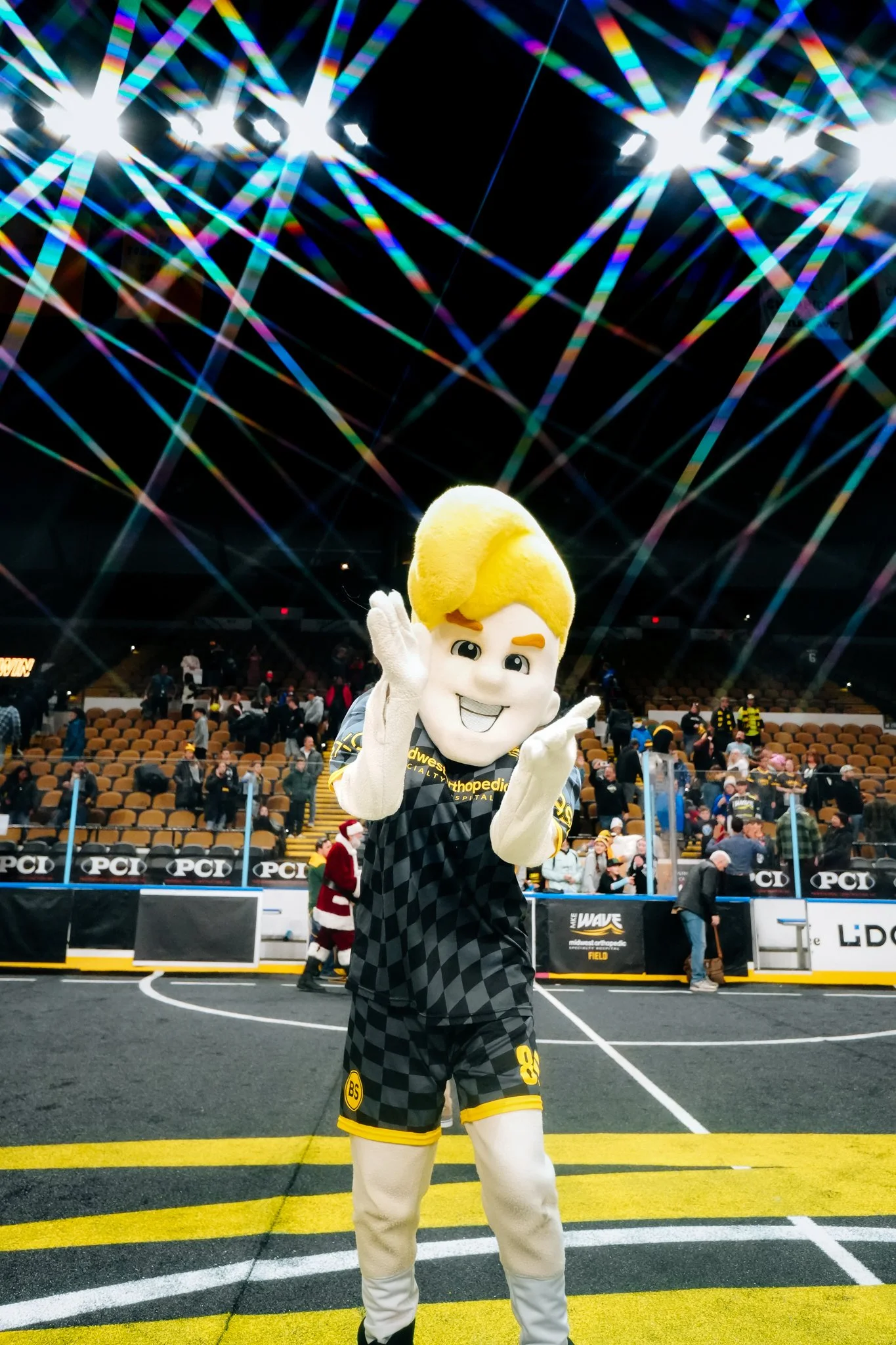 Mascot with blonde hair, wearing a black sports uniform with yellow accents, posing and smiling on a sports field with a crowd in the background and bright stadium lights overhead.