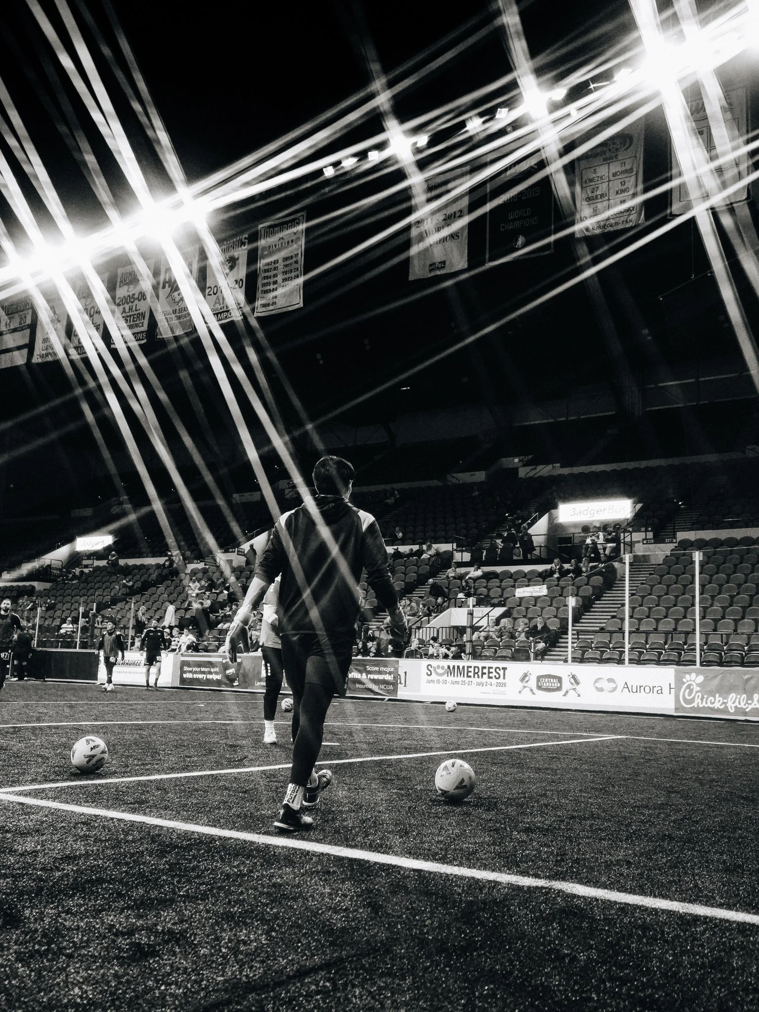 A person on a soccer field preparing for practice or game, with soccer balls on the ground and empty stadium seats in the background, under bright stadium lights in black and white.