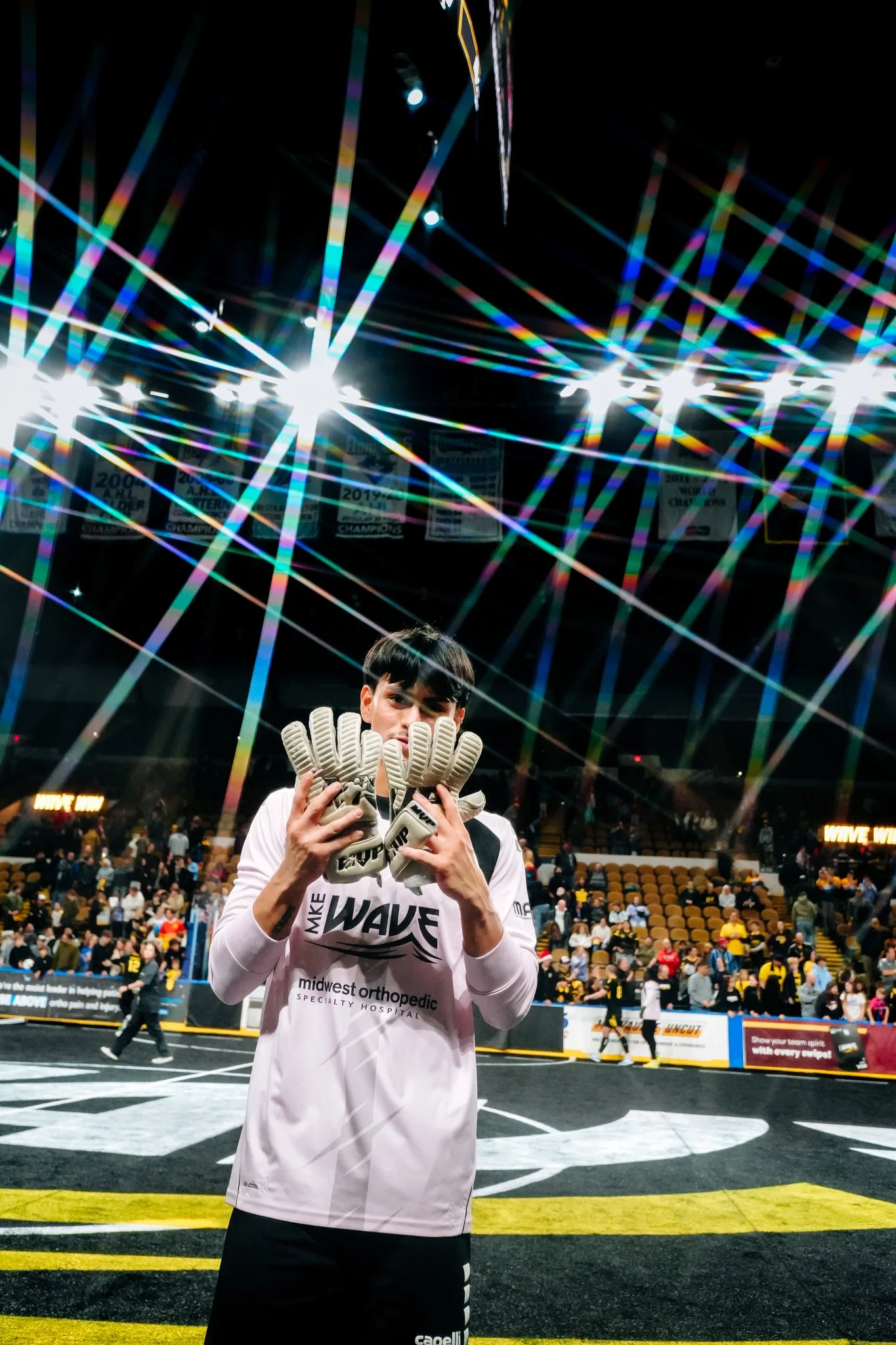 A young male athlete holding a pair of goalkeeper gloves on a sports field inside an arena, with spectators in the background and championship banners hanging from the ceiling.