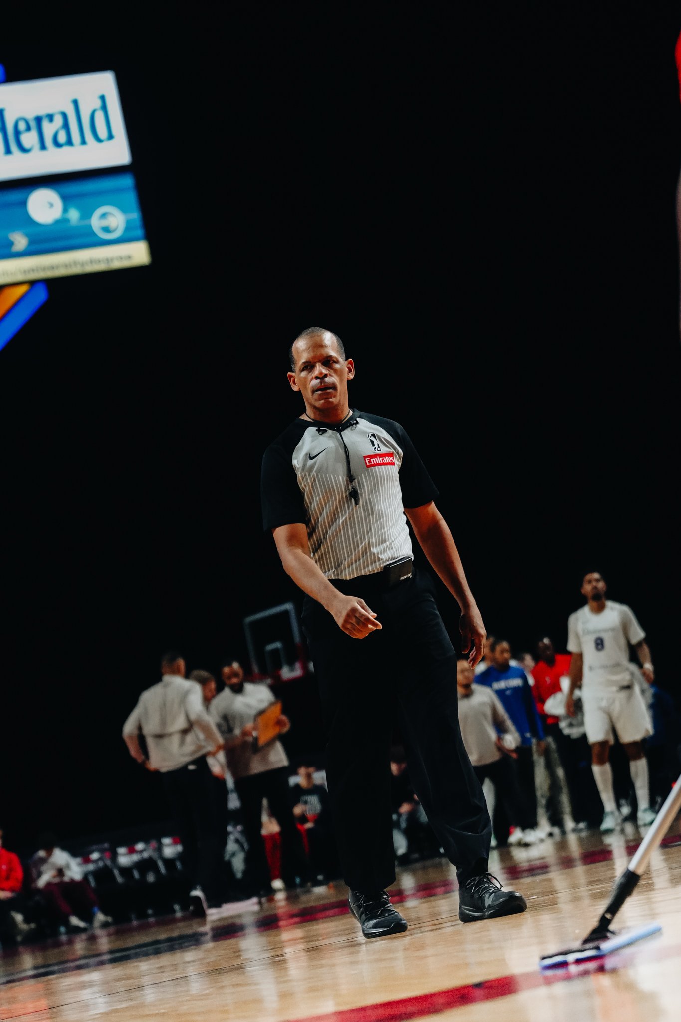 Basketball referee standing on the court during a game, with players and staff in the background.