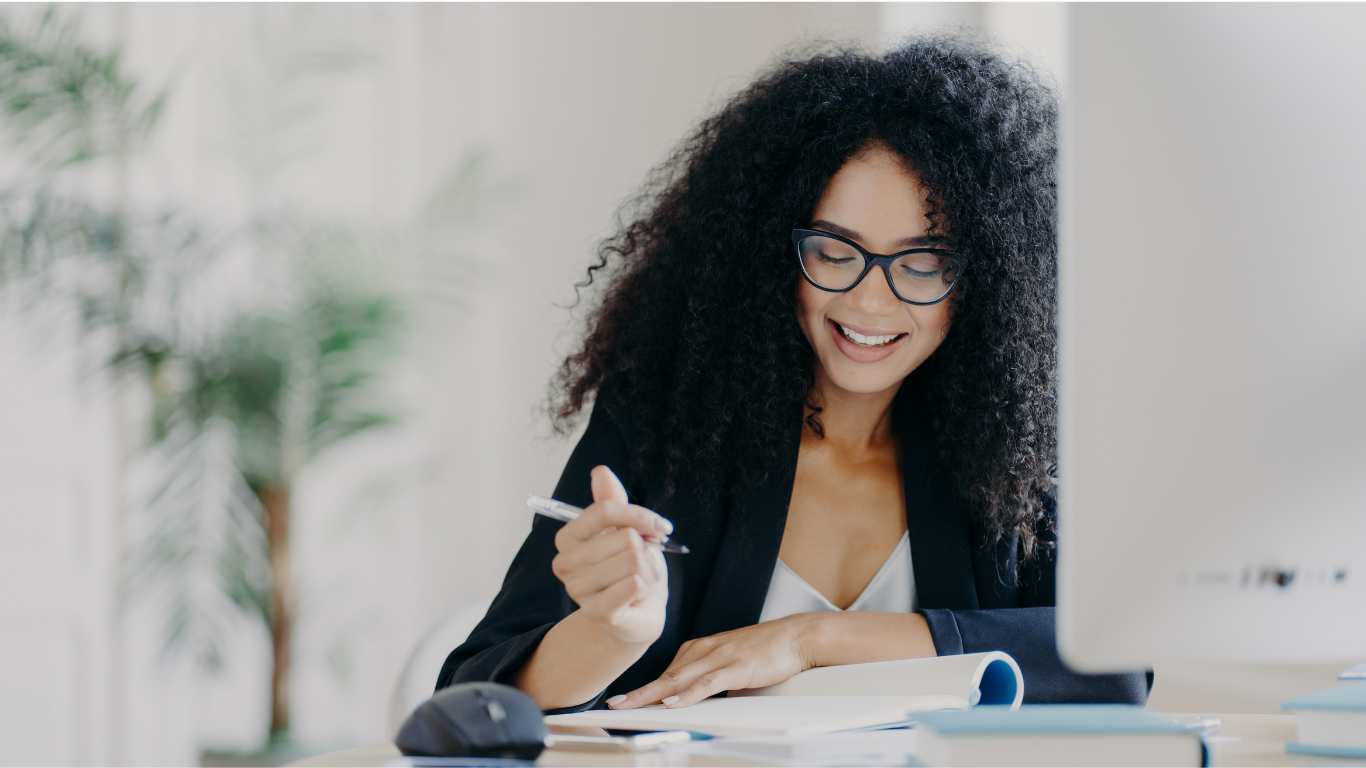A woman with curly black hair and glasses smiling while working at her desk with a pen and a computer in front of her.