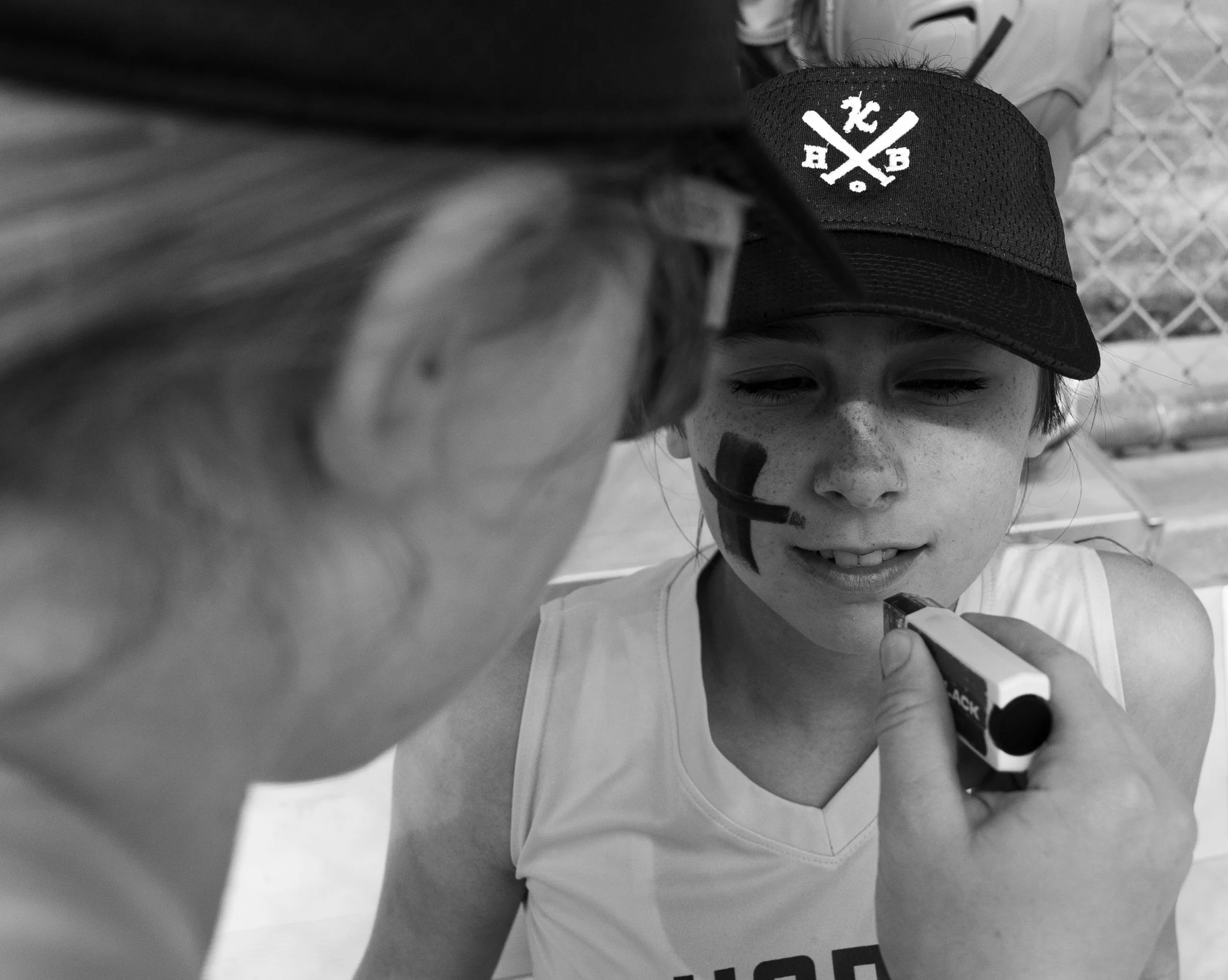A young girl sitting outdoors with face paint being applied by another person. The girl is wearing a baseball cap and a sleeveless shirt, and has her eyes partially closed, smiling slightly.