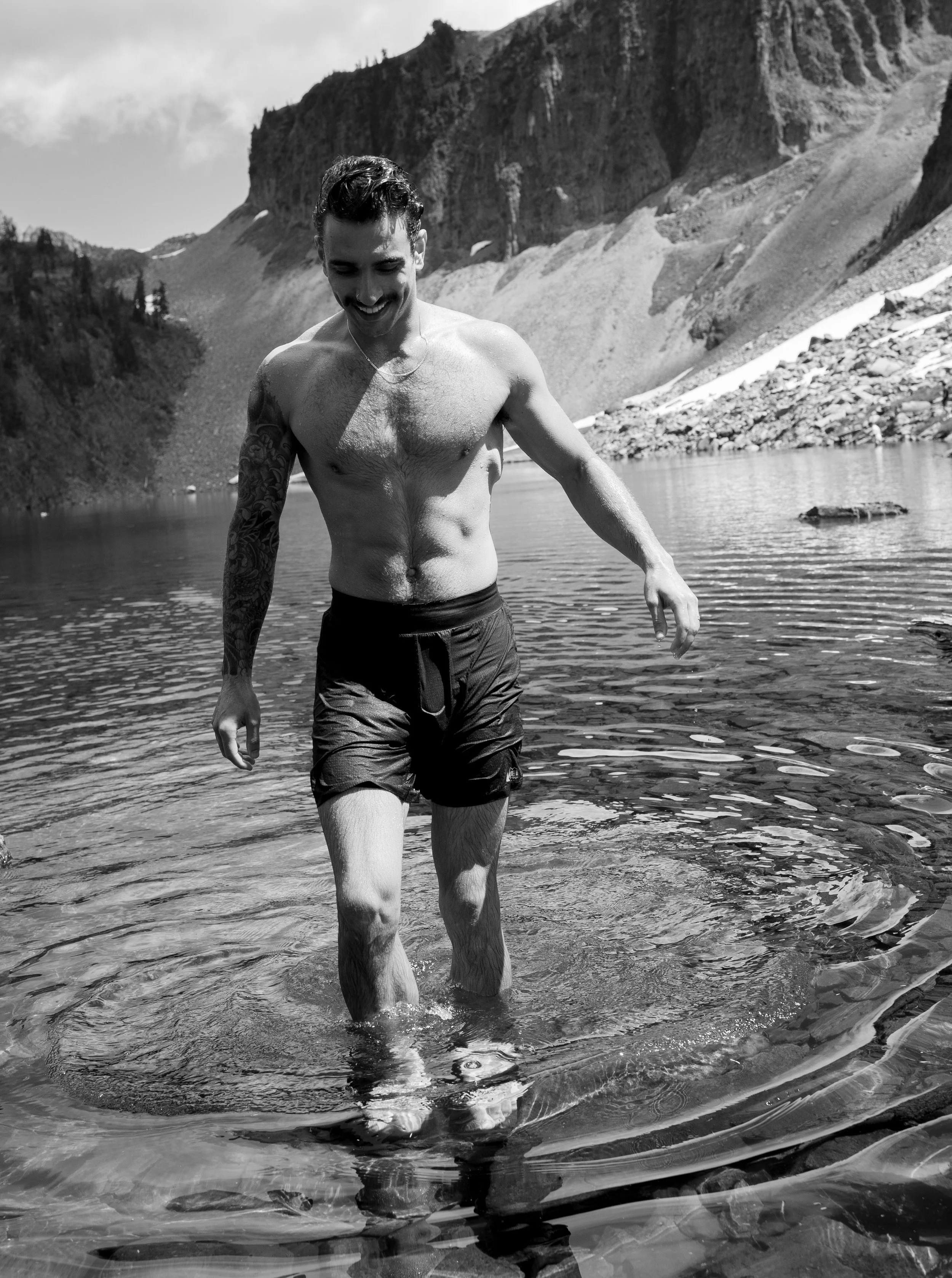 A shirtless man with a tattoo on his right arm, wearing black shorts, is smiling as he wades in a lake with a mountainous landscape in the background.