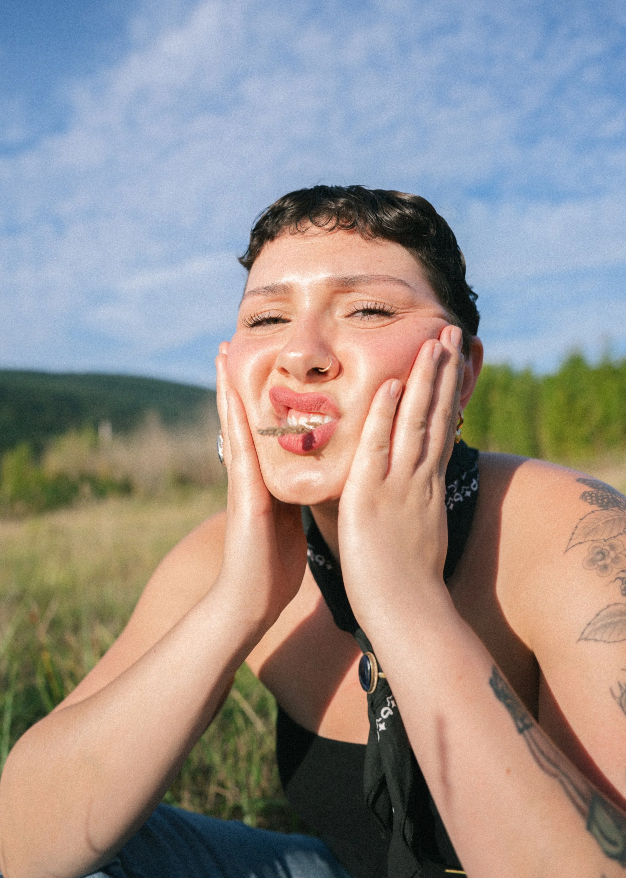 A person with short dark hair and tattoos, squinting with a playful expression, outdoors on a sunny day with blue sky and green landscape in the background.