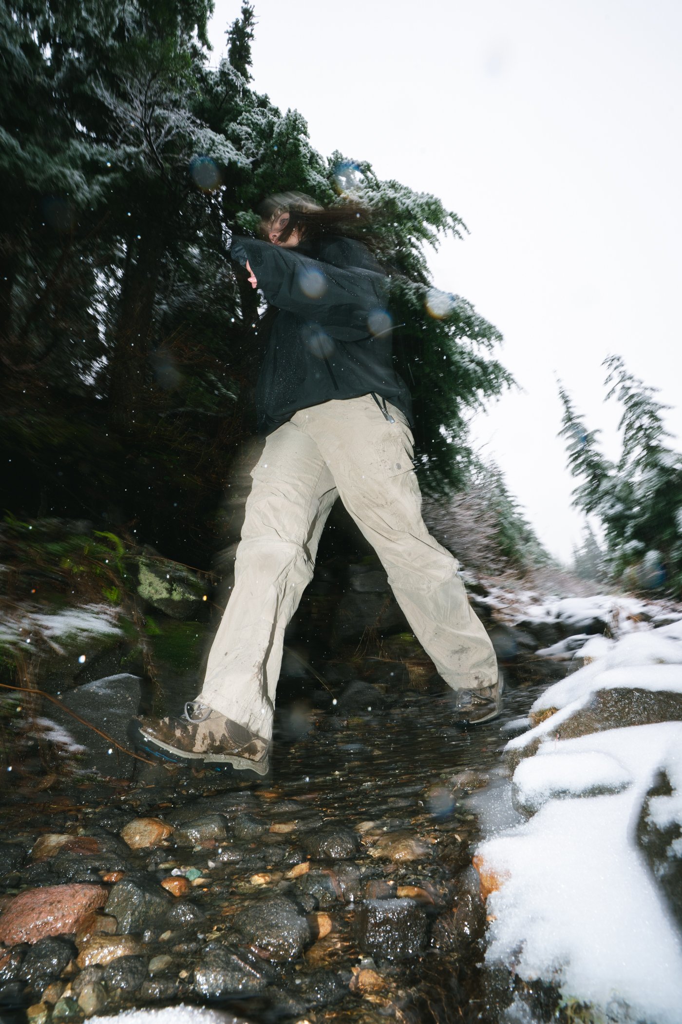 A person with long hair wearing a black jacket and light-colored pants walking over a rocky, snow-covered creek in a forested area during winter.