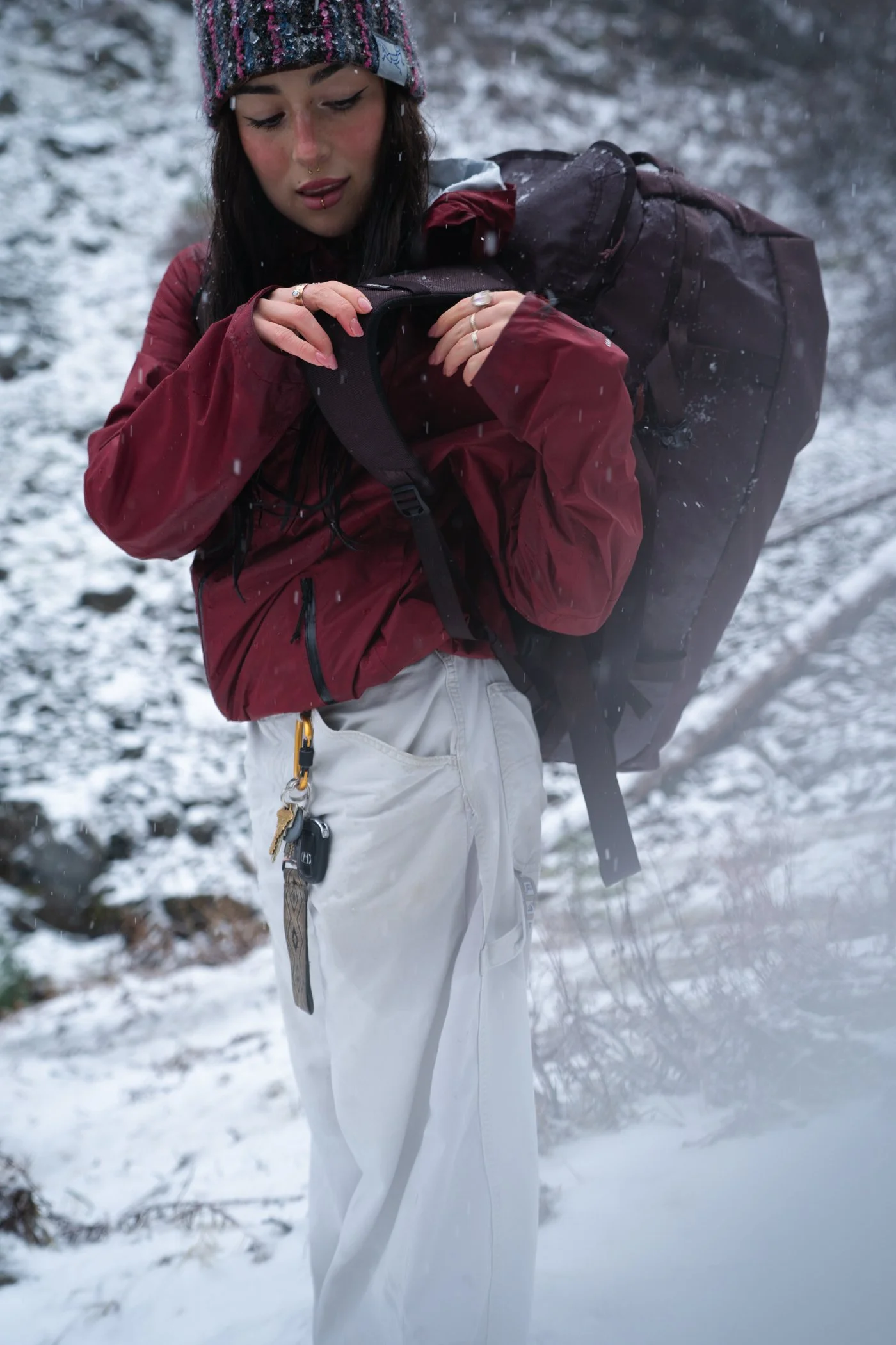 A woman outdoors in a snowy environment, wearing a red jacket, white pants, and a multicolored knit hat, adjusting her backpack strap.