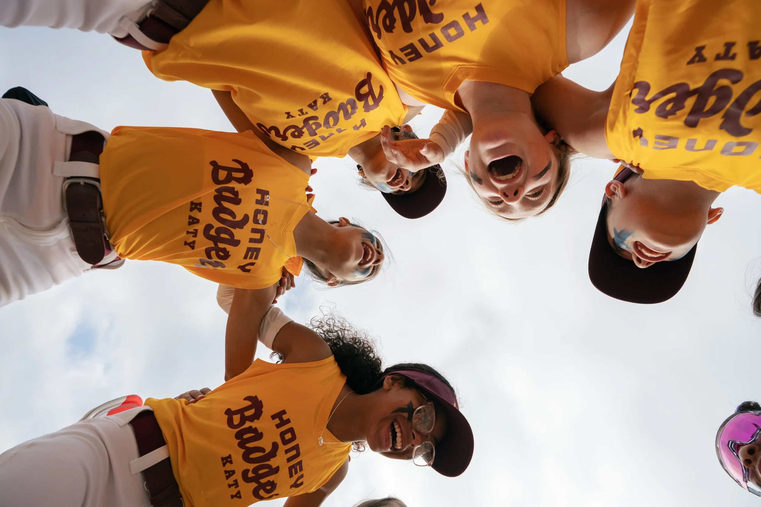 A softball team of young women wearing yellow shirts group together in a cheer before the game.