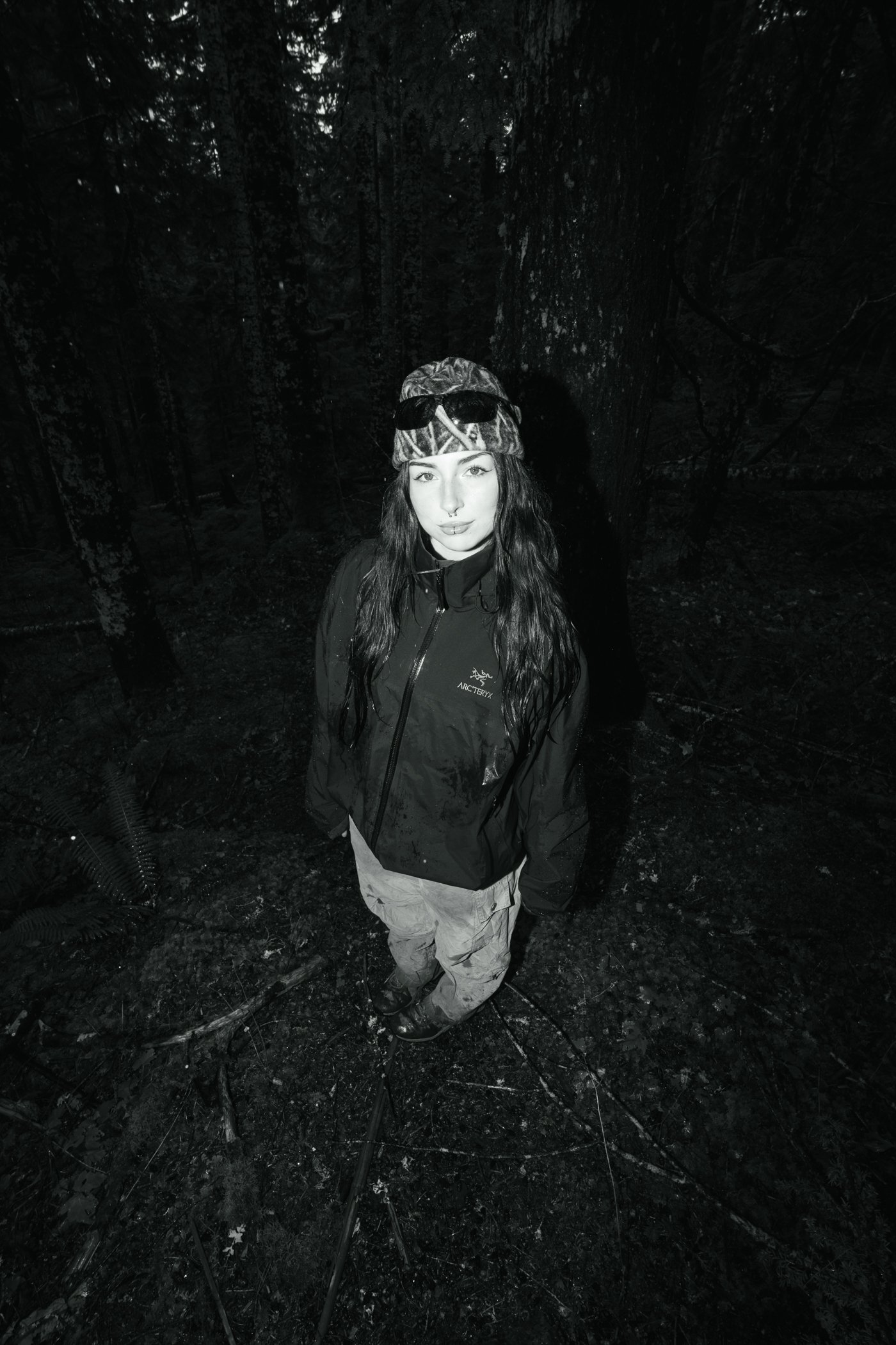 A woman standing in a dark forest wearing a raincoat and sunglasses with her dark hair down.