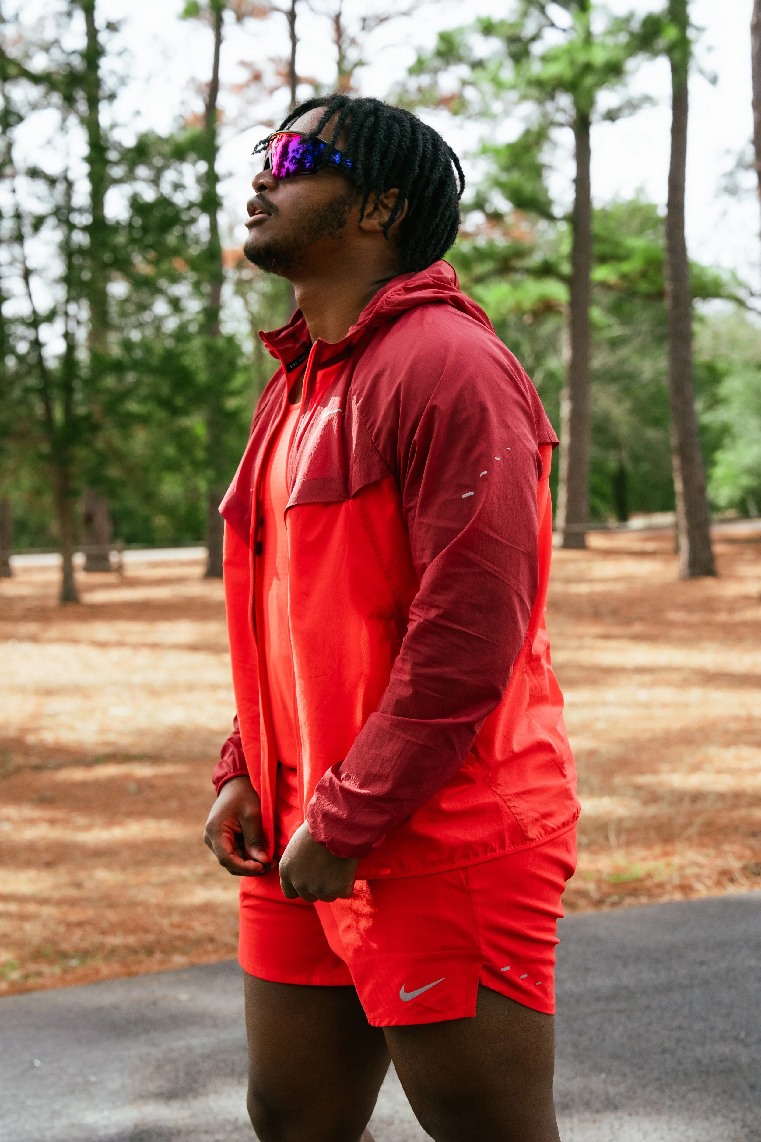 A man with dreadlocks and sunglasses in a red running jacket and shorts standing outdoors in a wooded park.