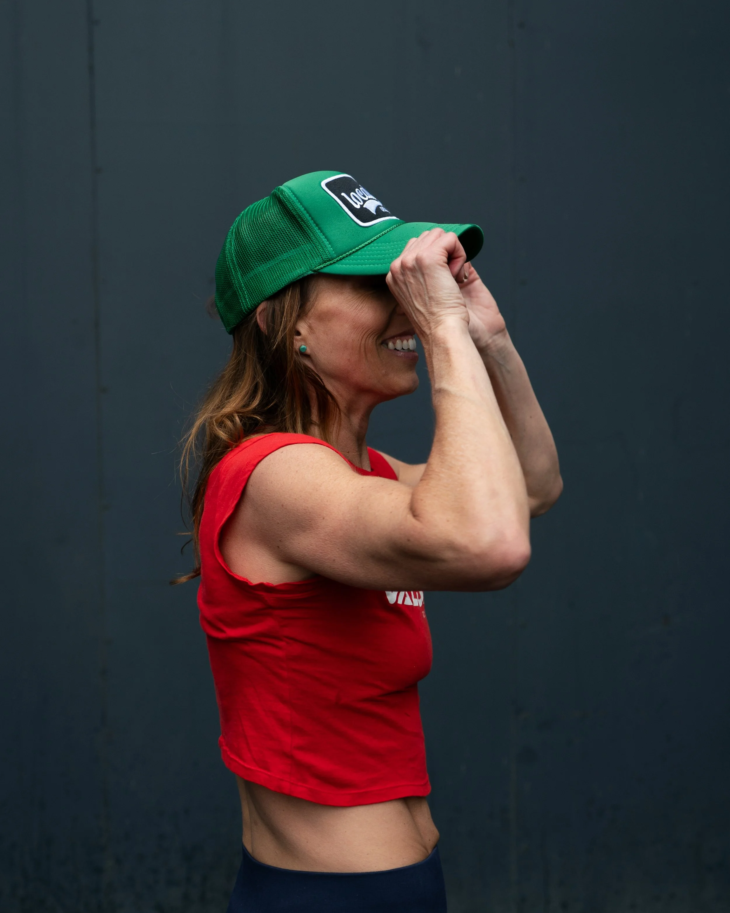 A smiling woman wearing a red sleeveless top and a green trucker hat is adjusting the hat with both hands, standing against a plain dark background.