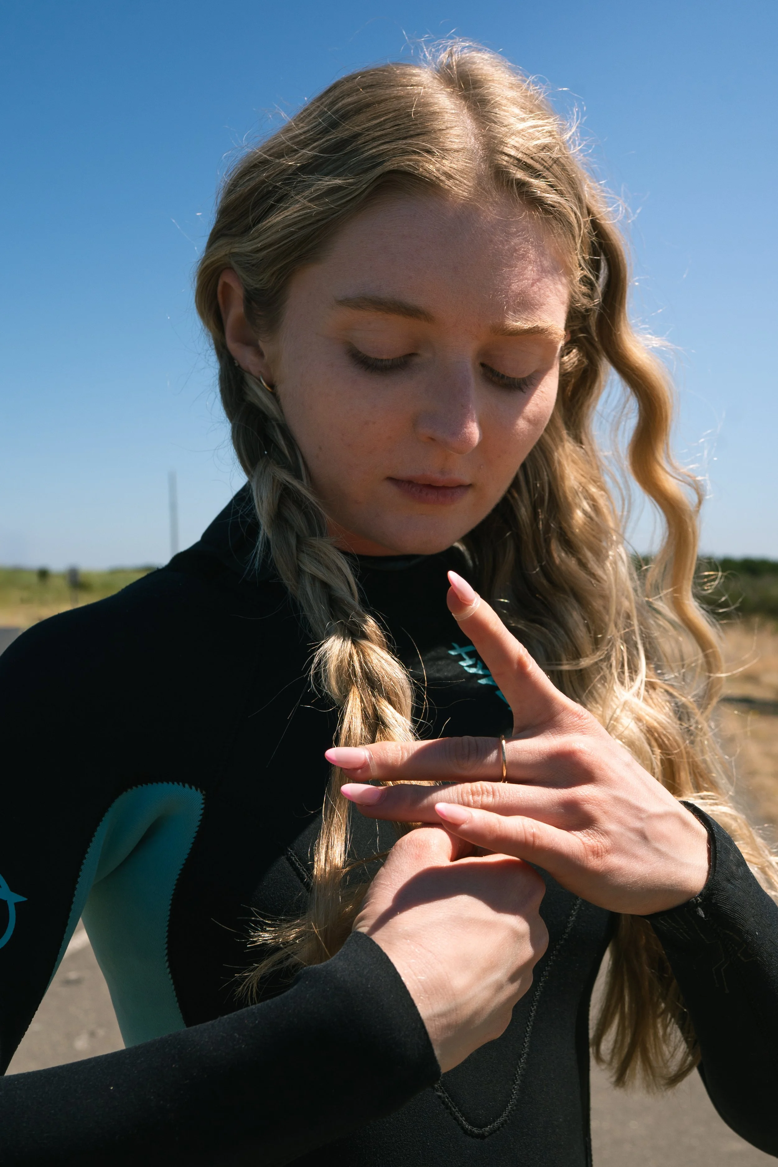 A young woman with long, wavy blonde hair puts hairbands in her braided hair on a sunny day.