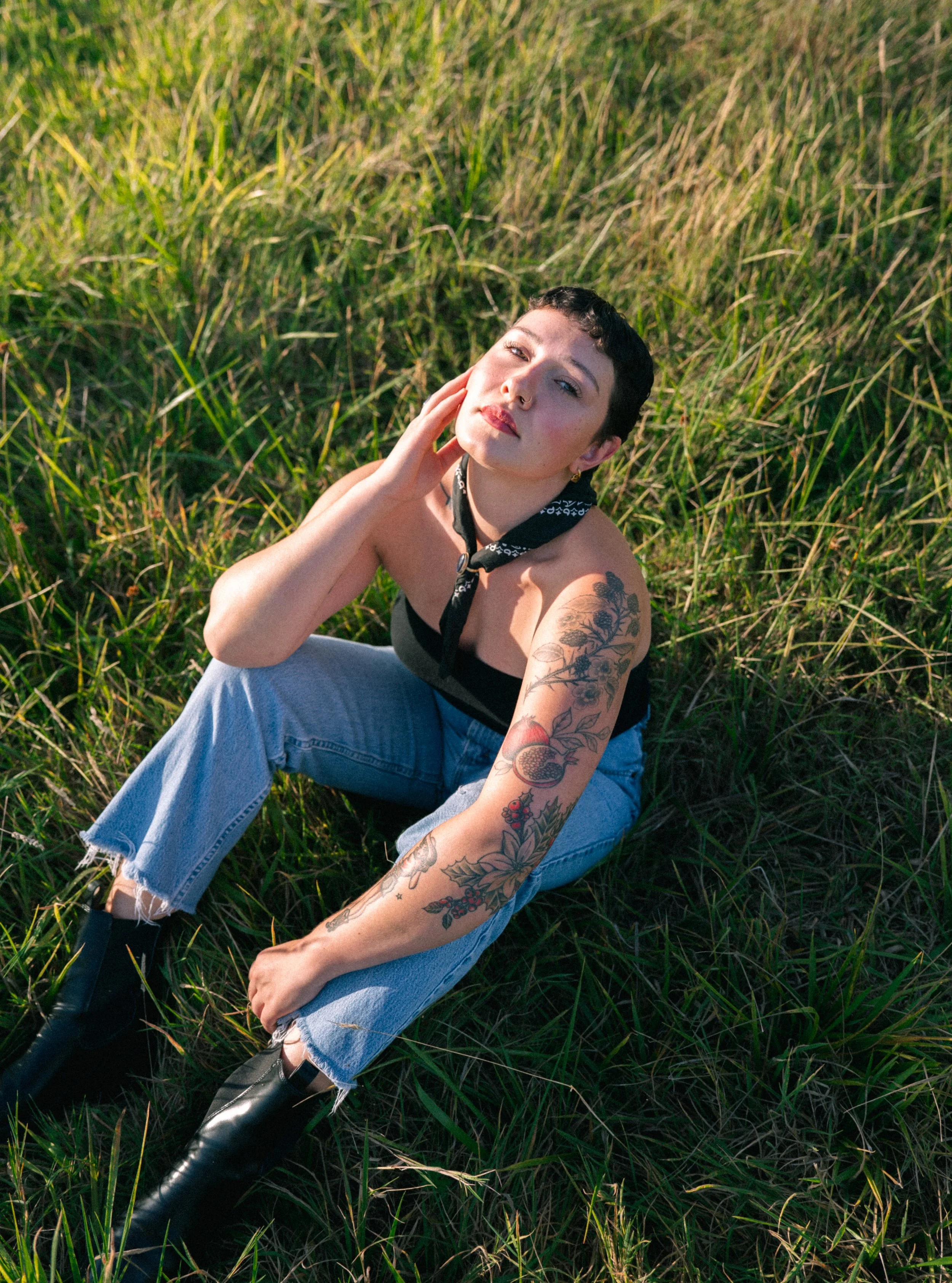 Young woman with short dark hair, tattoos on her left arm, wearing a black tube top, light blue ripped jeans, black boots, and a bandana around her neck, sitting on grass in a field, looking at the camera.