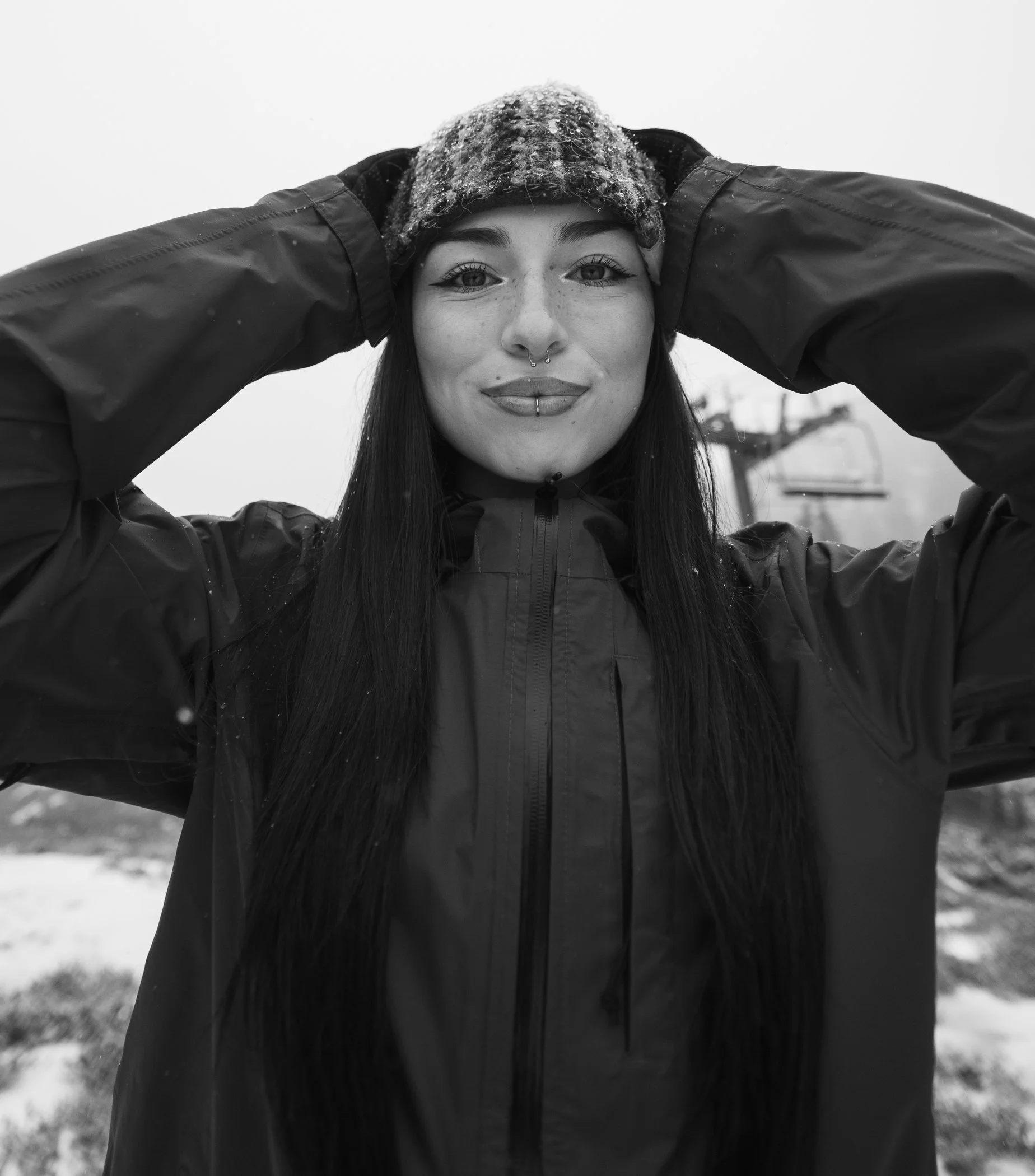 Black and white photo of a woman wearing a winter jacket and a beanie, holding her head with both hands, outdoors in snowy weather, with ski lift visible in the background.