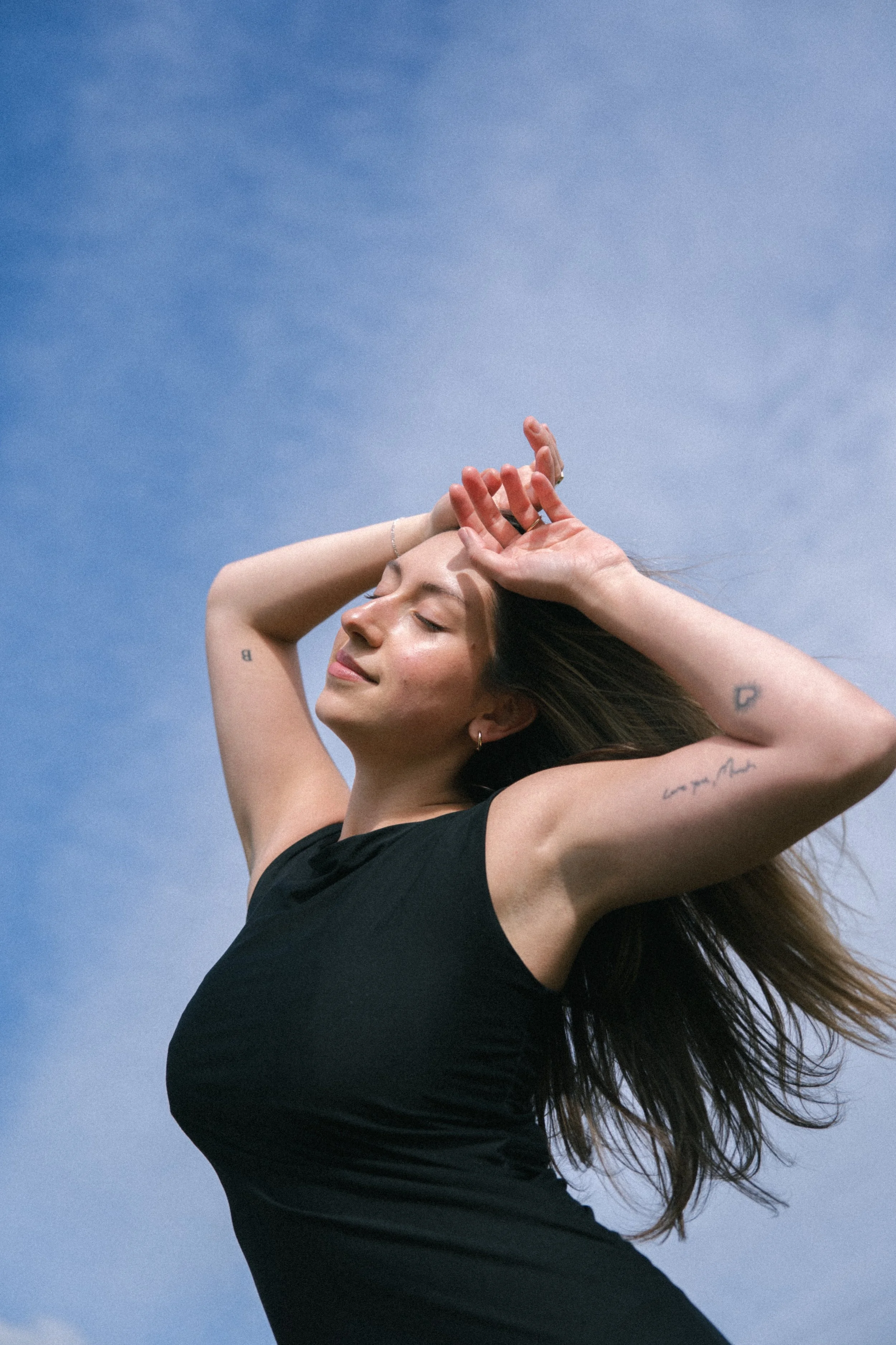 A woman with long hair wearing a black sleeveless shirt, standing outdoors with her eyes closed, smiling, and her hands on her head against a blue sky with wispy clouds.